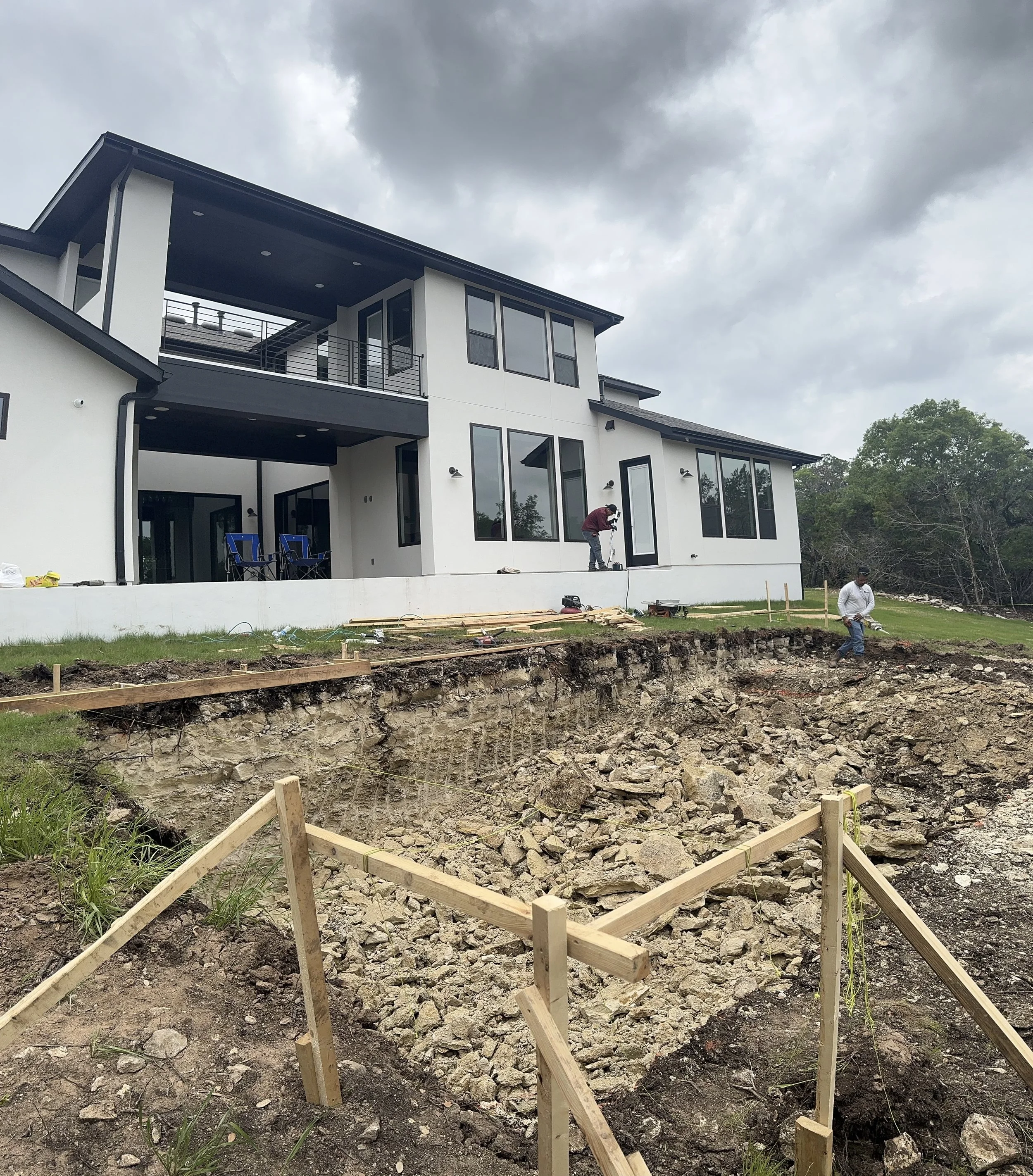 Construction workers working on a site in front of a modern house with large windows and an elevated porch, as the ground is excavated for foundation work.