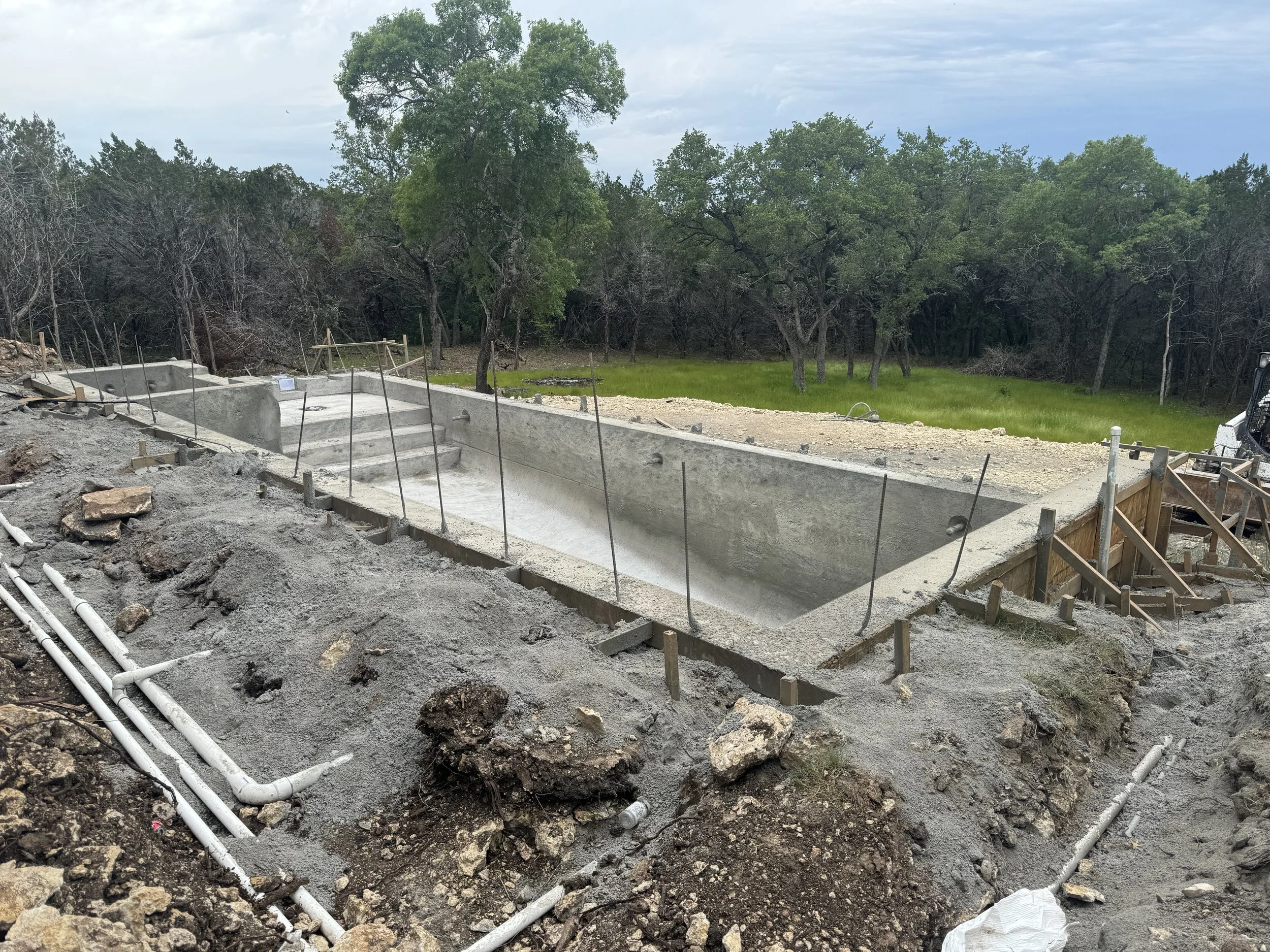 Under-construction swimming pool in backyard with concrete stairs, surrounded by dirt, construction materials, and scaffoldings, with trees and grassy area in background.
