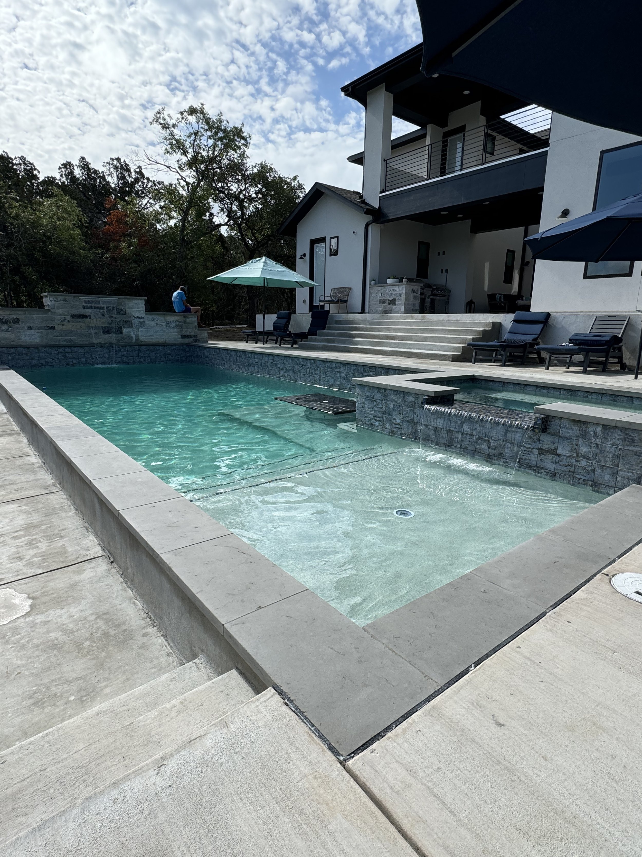 Modern backyard with a swimming pool, poolside chairs, and house with two floors, umbrellas, and outdoor furniture under a cloudy sky.