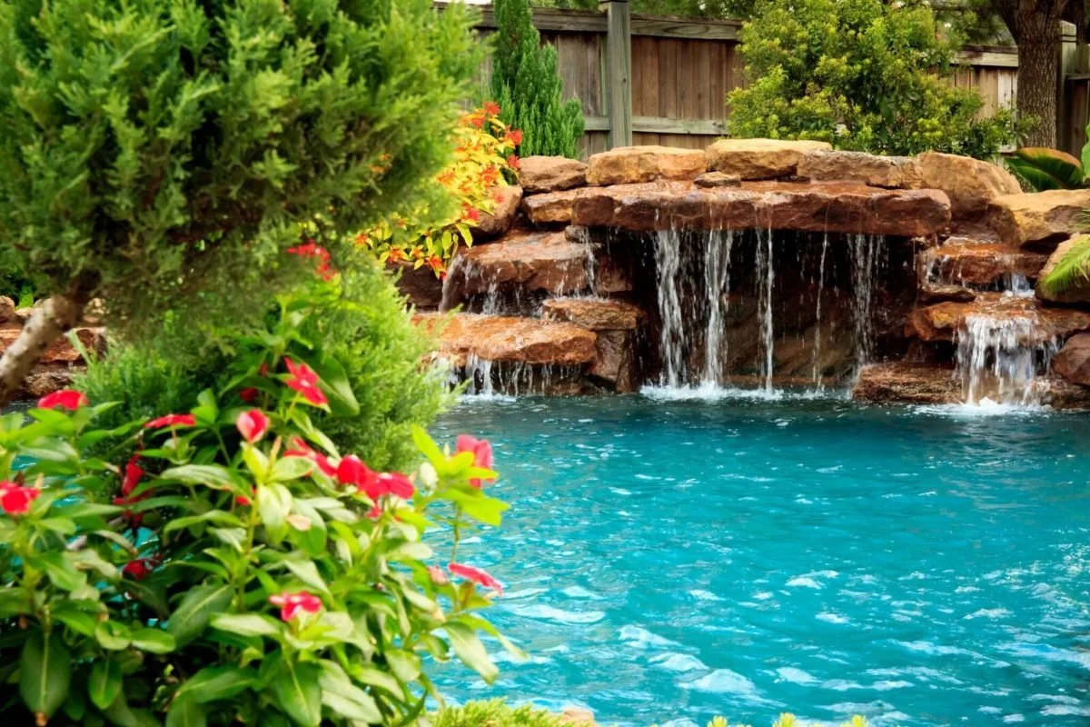 A backyard with a small waterfall and a swimming pool surrounded by lush green plants and flowers.