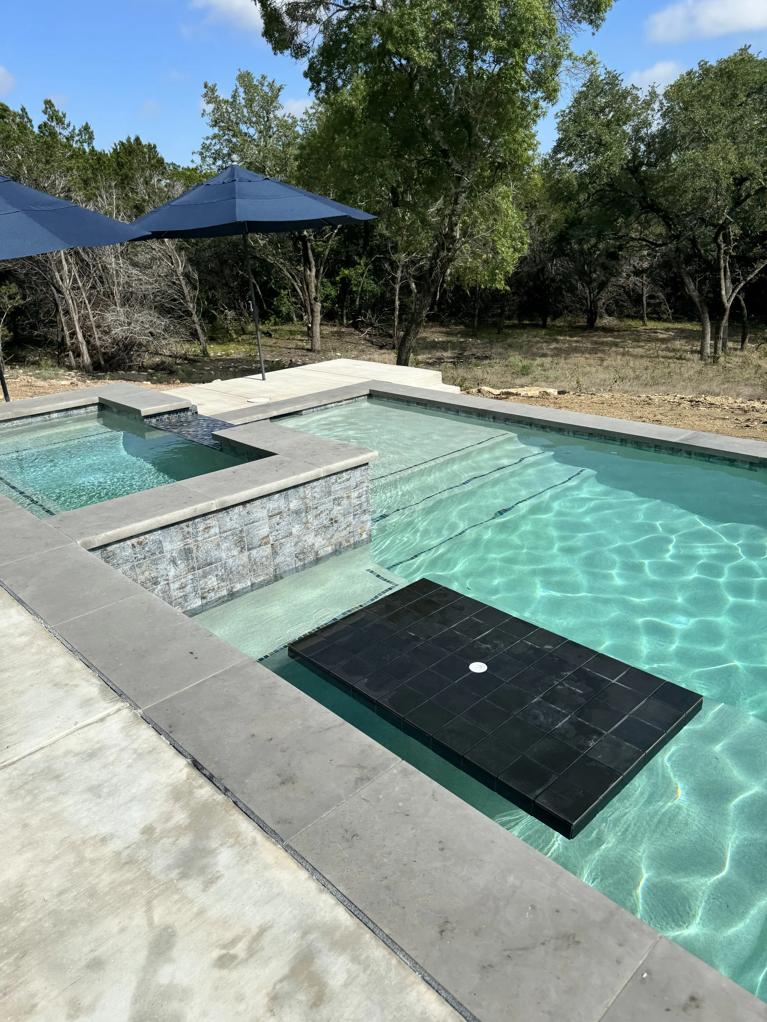 Luxury backyard swimming pool with a built-in hot tub, waterfall feature, and black tiled island, surrounded by trees and umbrellas.