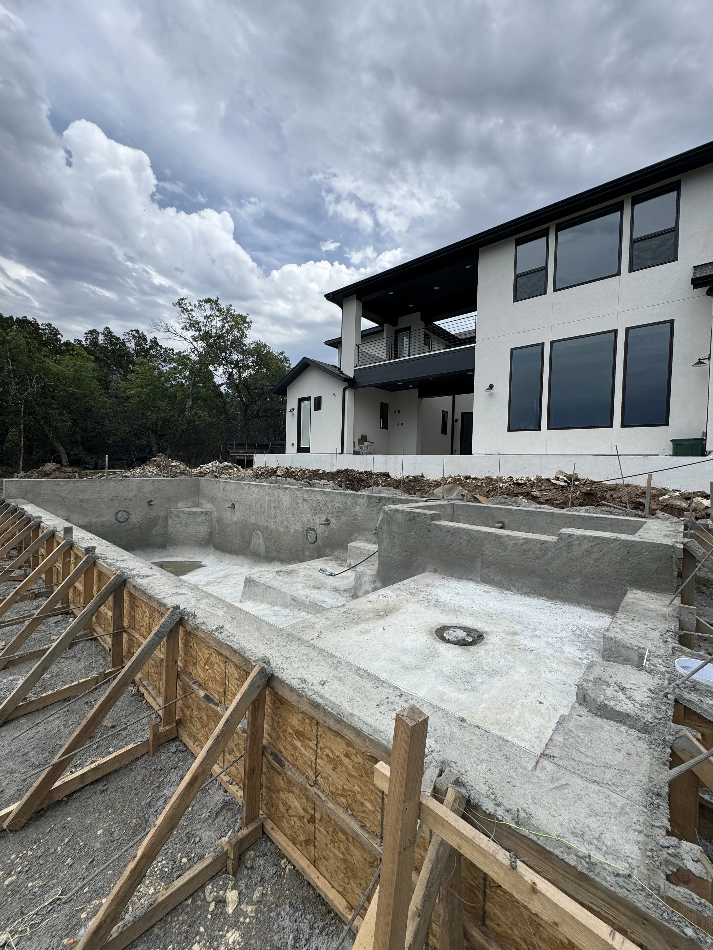 Construction site of a backyard swimming pool in front of a modern two-story house with large windows, with cloudy sky overhead.