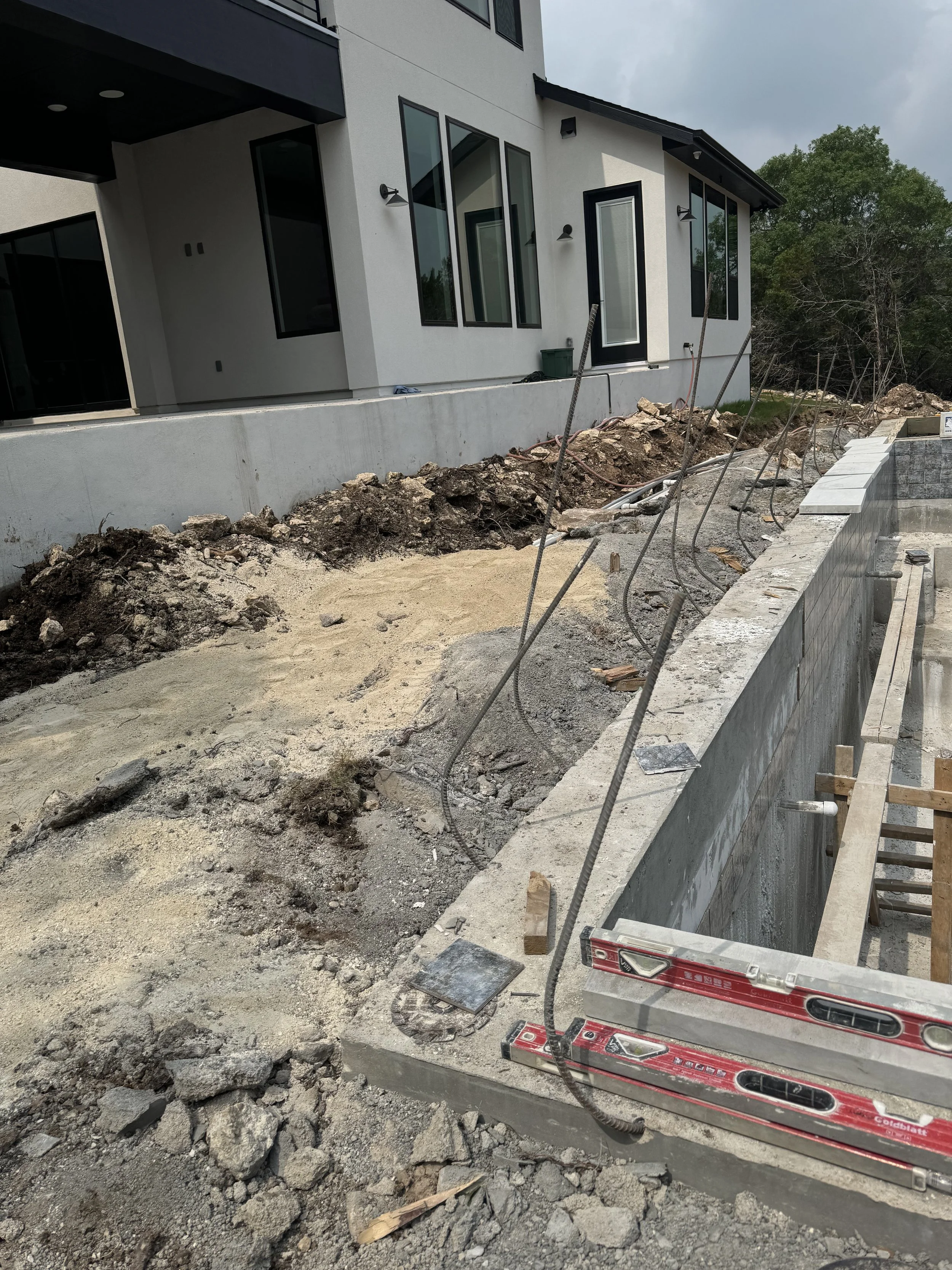 Construction site in the backyard of a modern house with a partially built pool or foundation area, exposing concrete and rebar, with construction tools and uneven dirt ground.