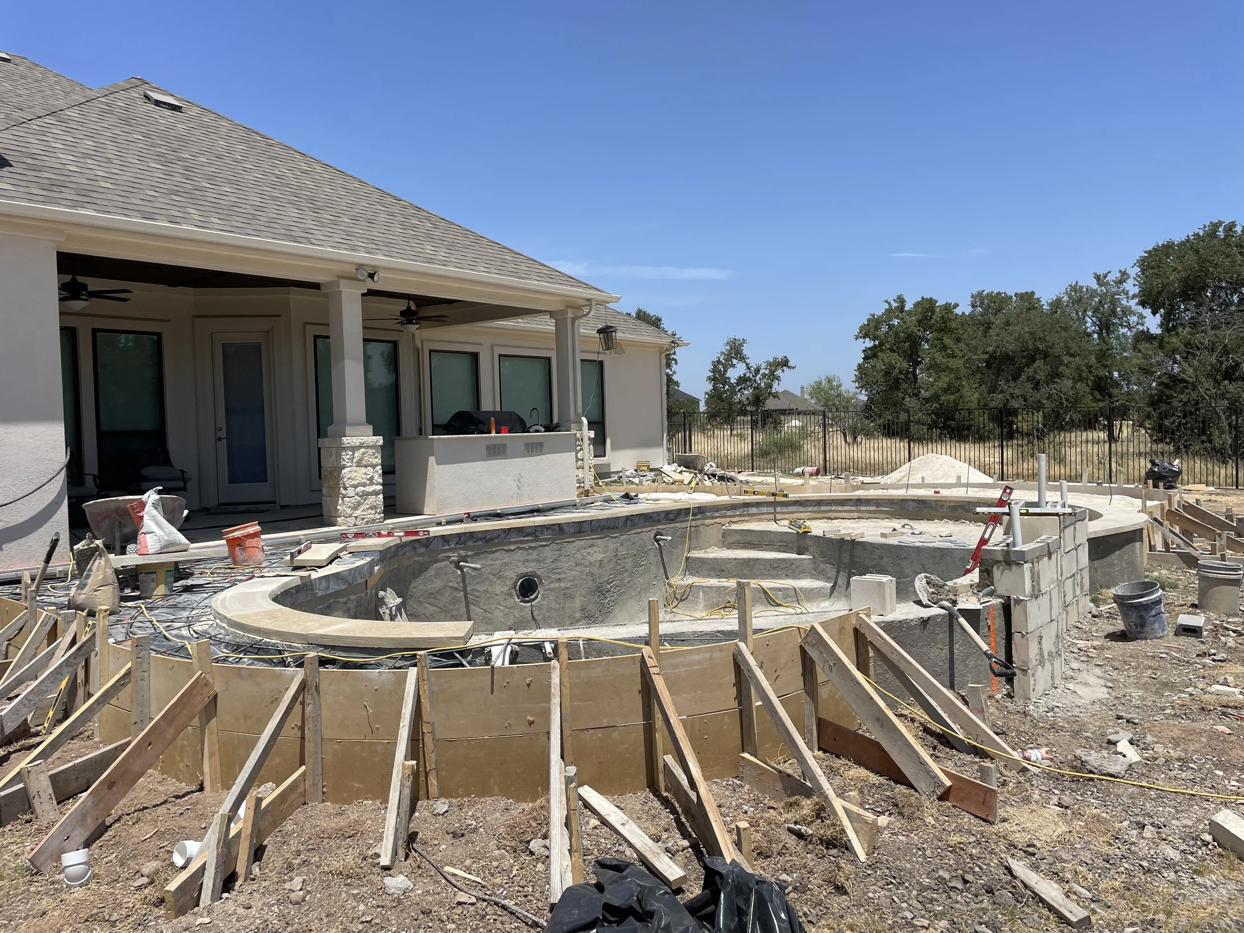 Construction site of a backyard swimming pool, with a house in the background, showing construction materials, equipment, and partial fencing.