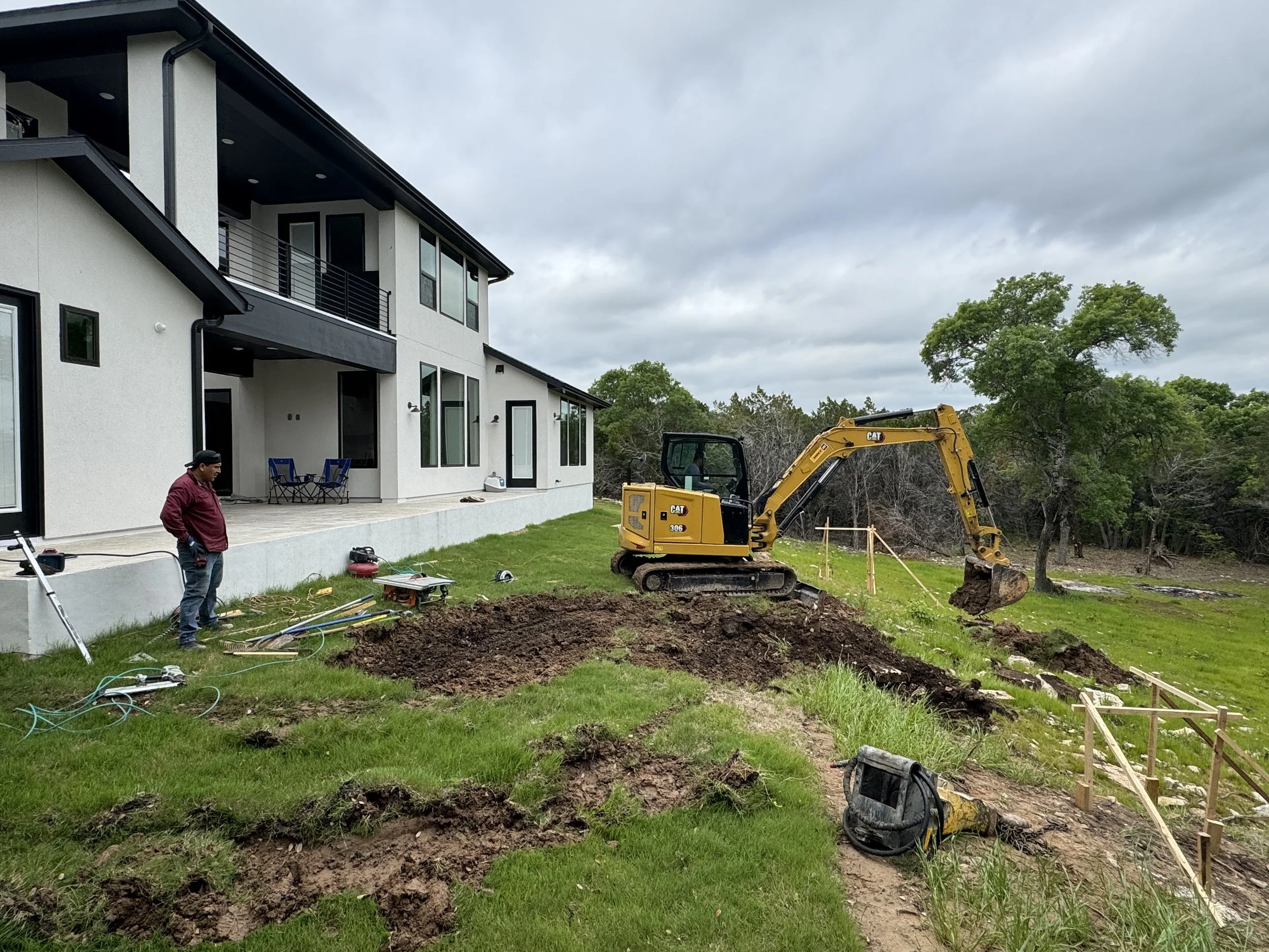 Construction workers and equipment excavating soil in backyard of a modern house, with trees and cloudy sky in background.