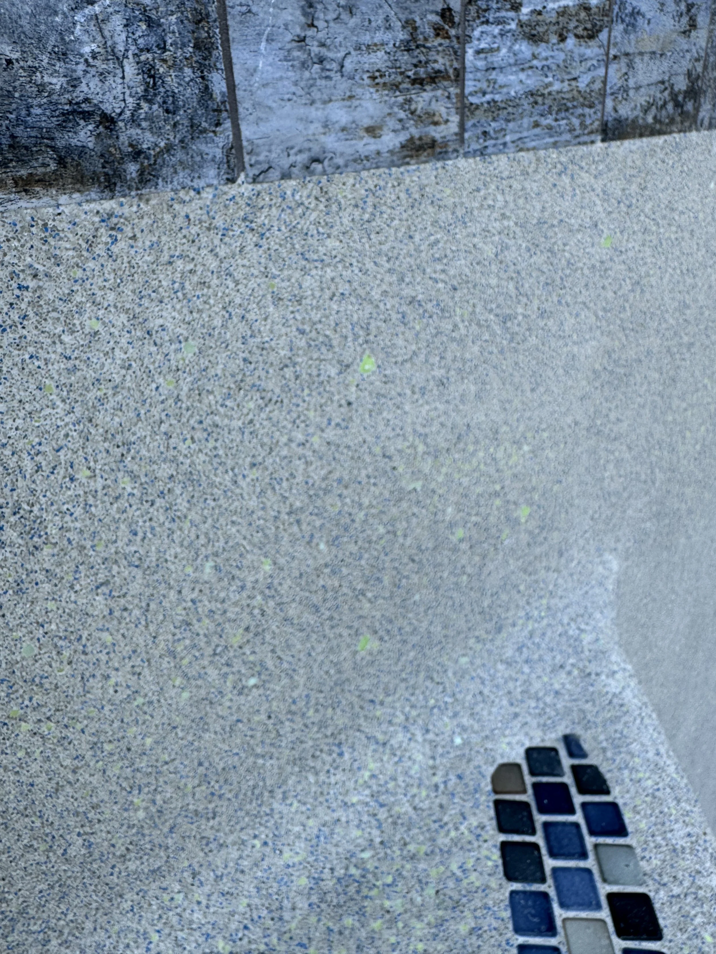 Close-up of a speckled grey surface, possibly a countertop or wall, with a small rectangular color tile mosaic near the bottom right corner.