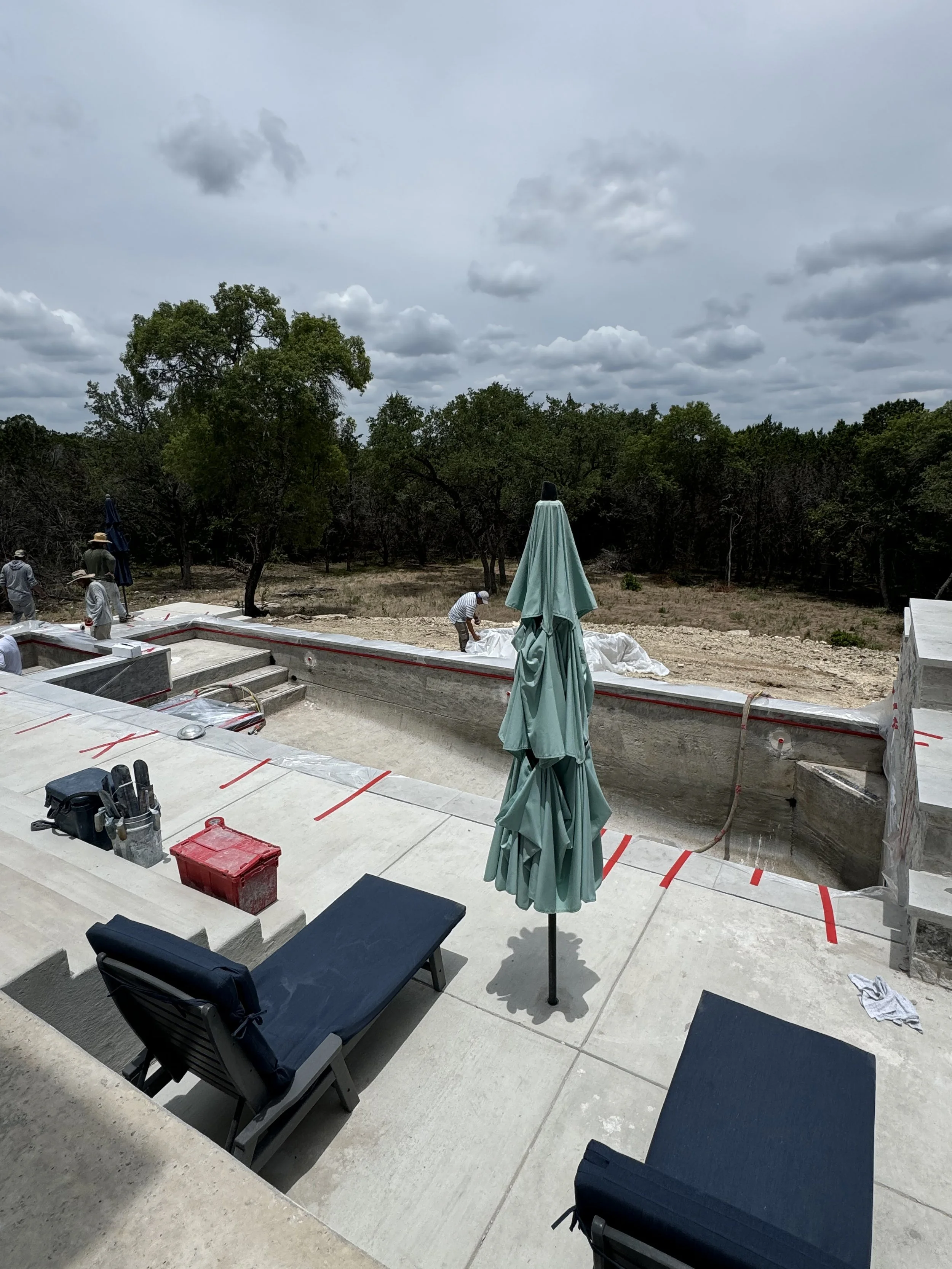 Construction workers working on a backyard swimming pool, with a patio area and lounge chairs, under a cloudy sky.