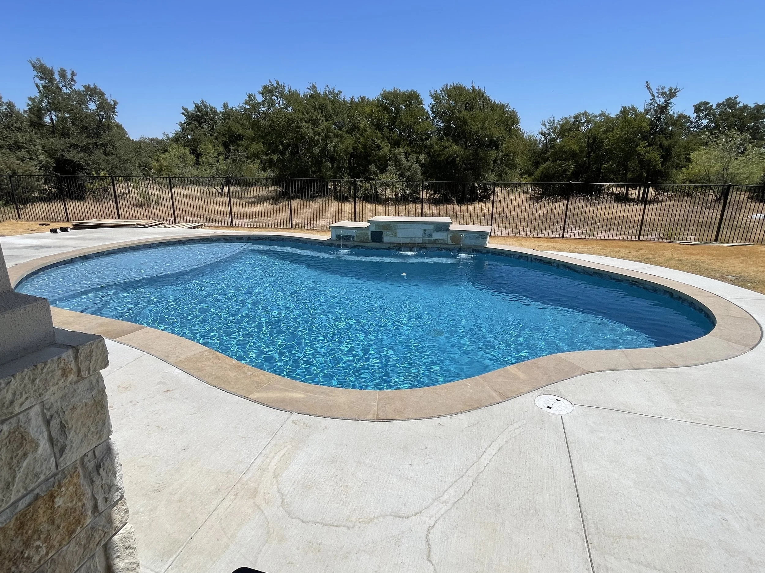 A backyard swimming pool with a curved shape surrounded by a concrete deck, a black metal fence, green trees, and a clear blue sky.