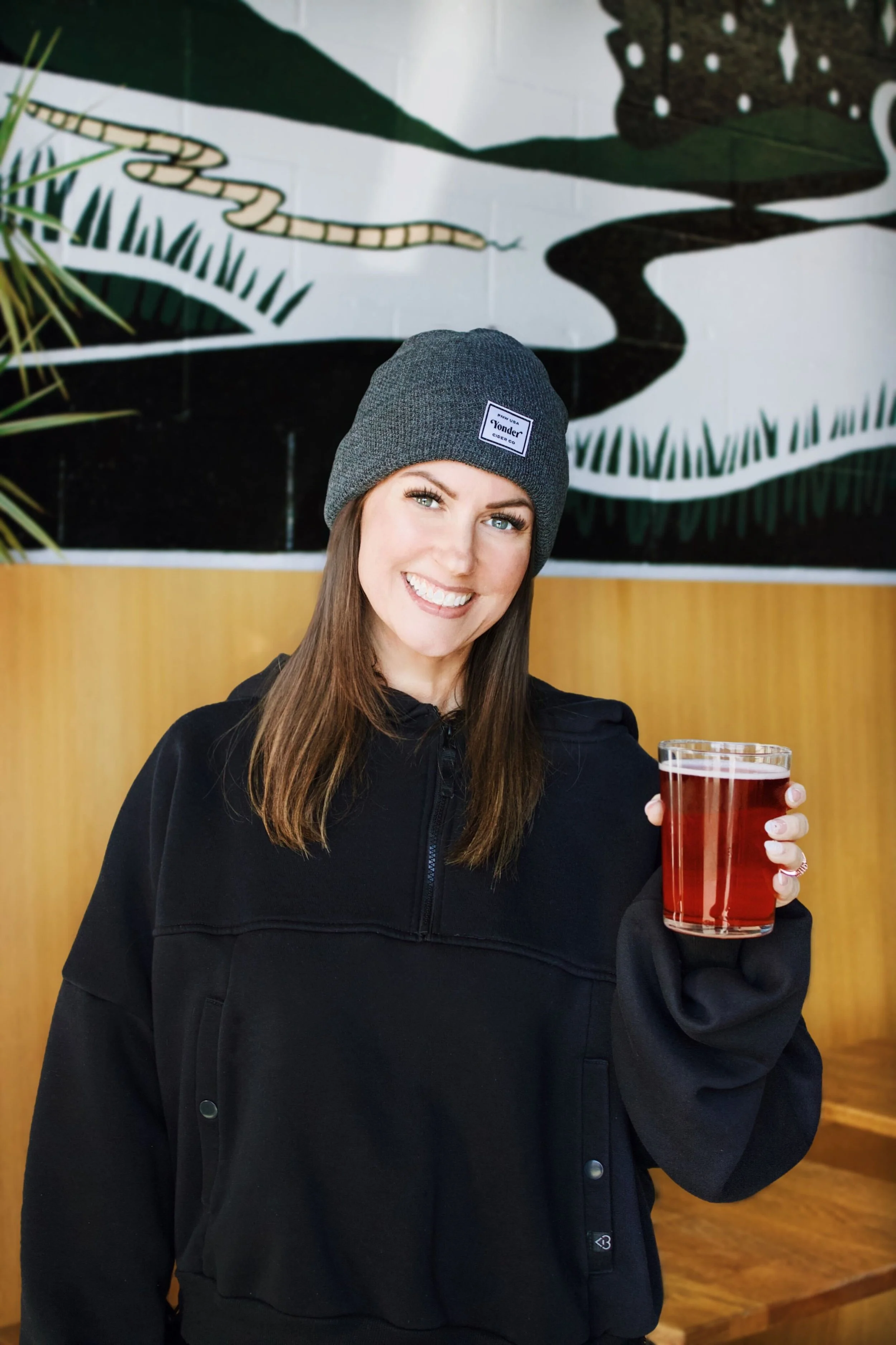 A woman in a green sweatshirt smiling and holding a glass of beer at a bar or taproom with a menu board behind her listing various drinks.