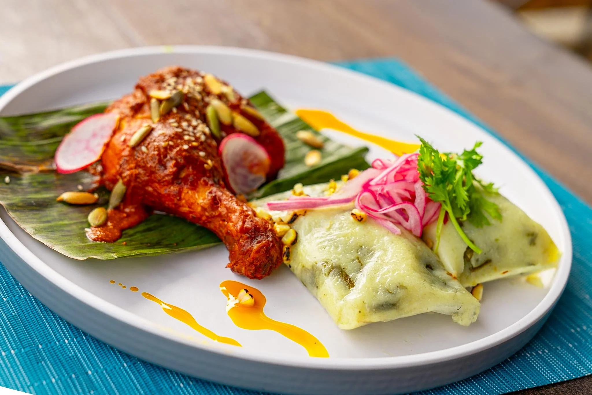 Plate with cooked chicken leg, radish slices, cabbage slices, cilantro, and green sauce, garnished with seeds and a drizzle of yellow sauce.