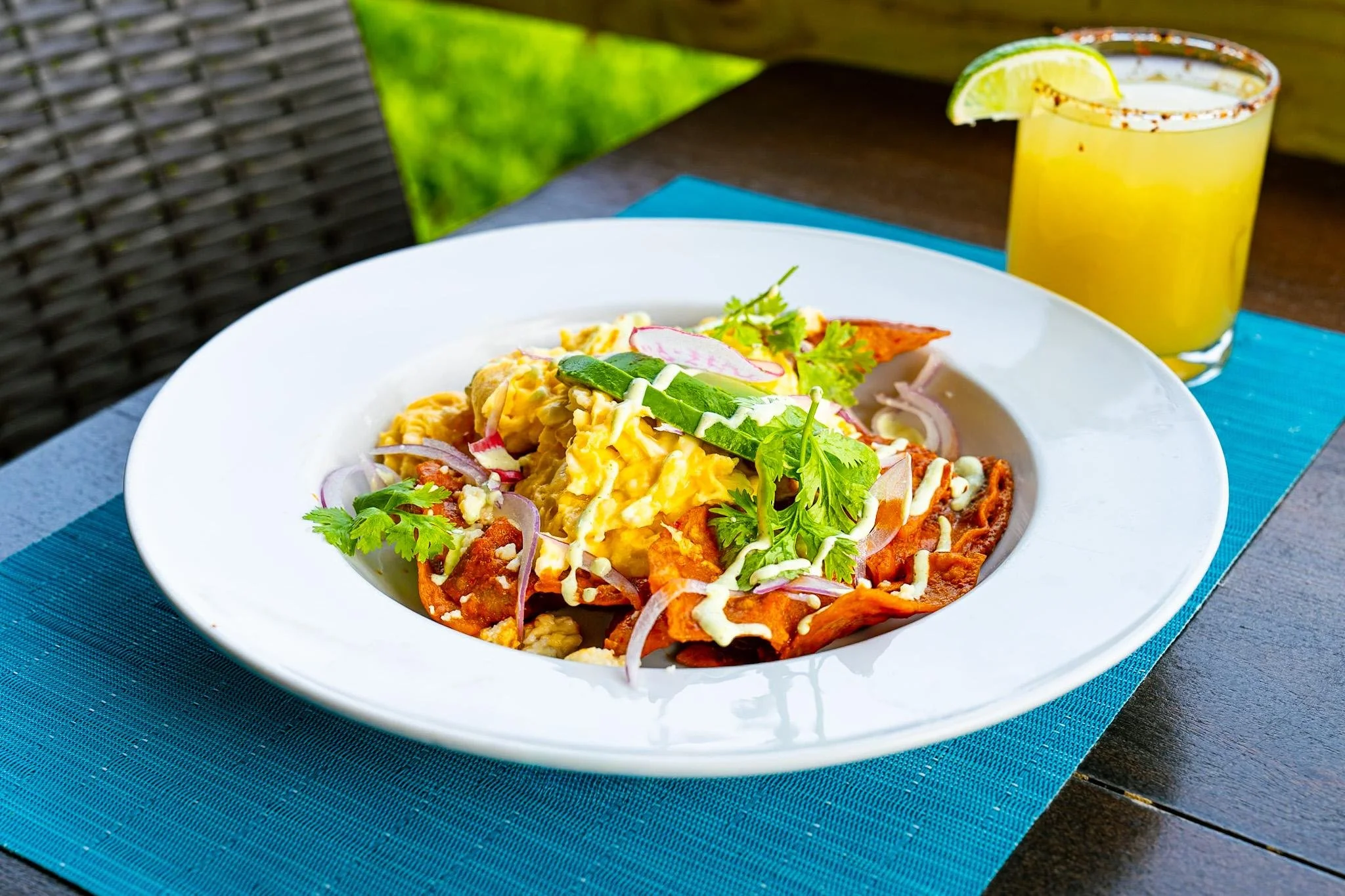 Plate of Mexican nachos topped with scrambled eggs, sliced radishes, a sprig of cilantro, and drizzled with sauce, with a lime wedge and yellow drink in the background.