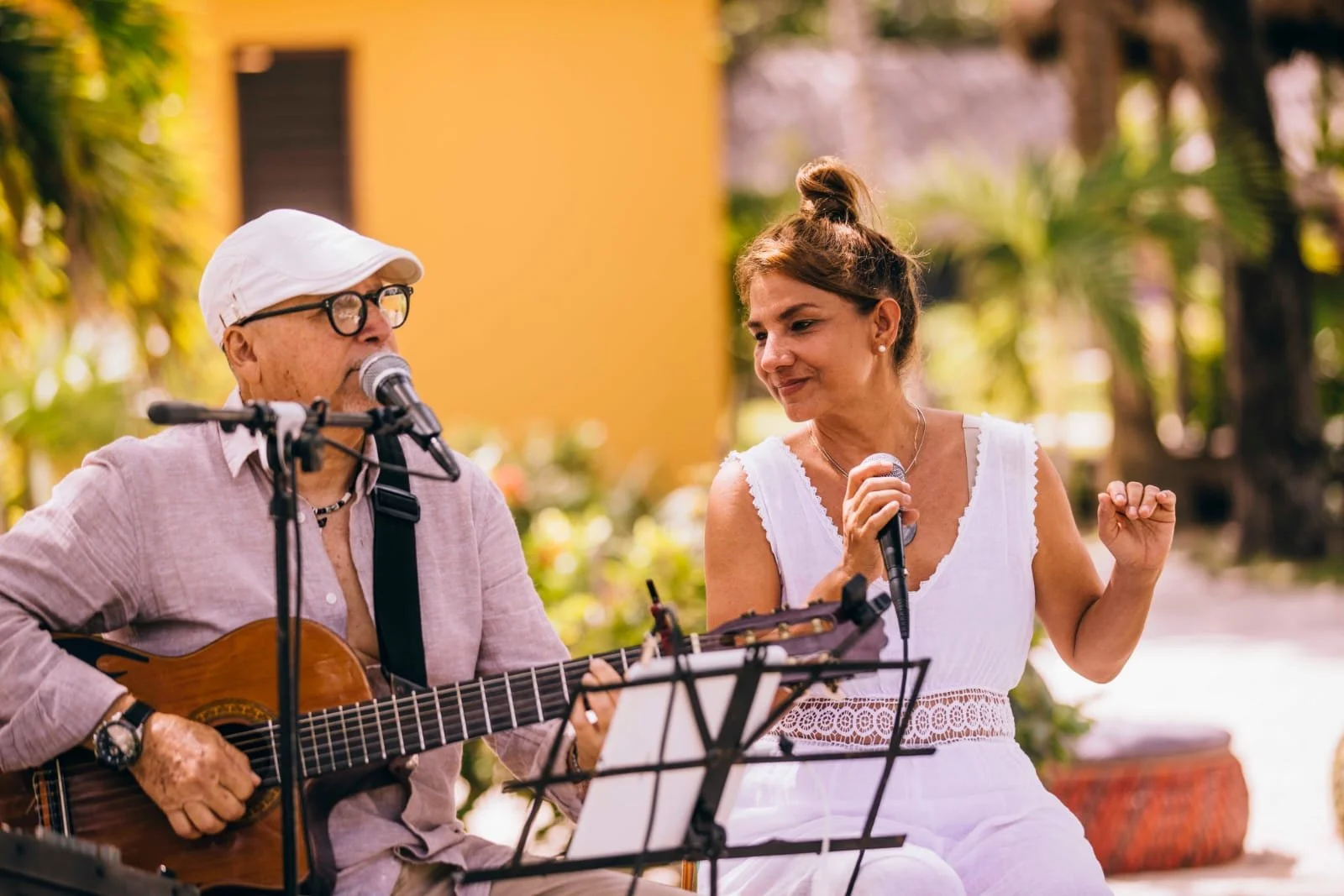 Older man playing guitar and singing into a microphone with a woman singing along, outdoors with green plants and a yellow building in the background.