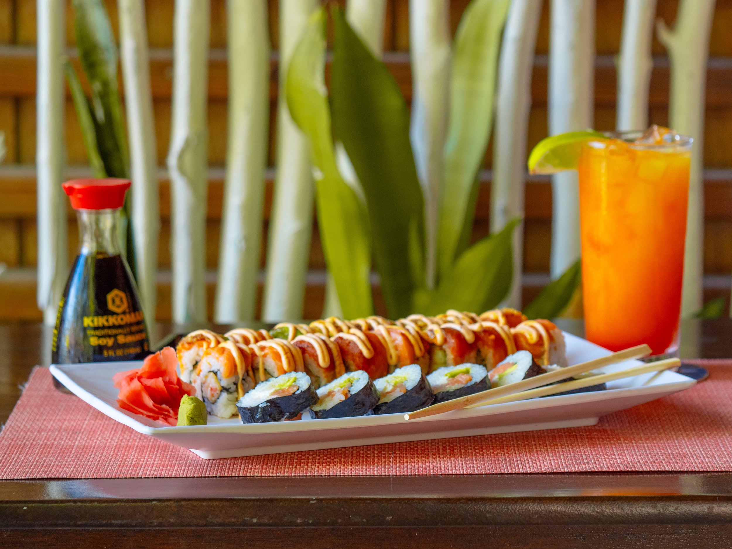 A plate of sushi rolls with various toppings, pickled ginger, and wasabi, accompanied by a tall glass of orange drink with a lime wedge and a bottle of soy sauce on a wooden table with a red placemat, green plants, and bamboo in the background.
