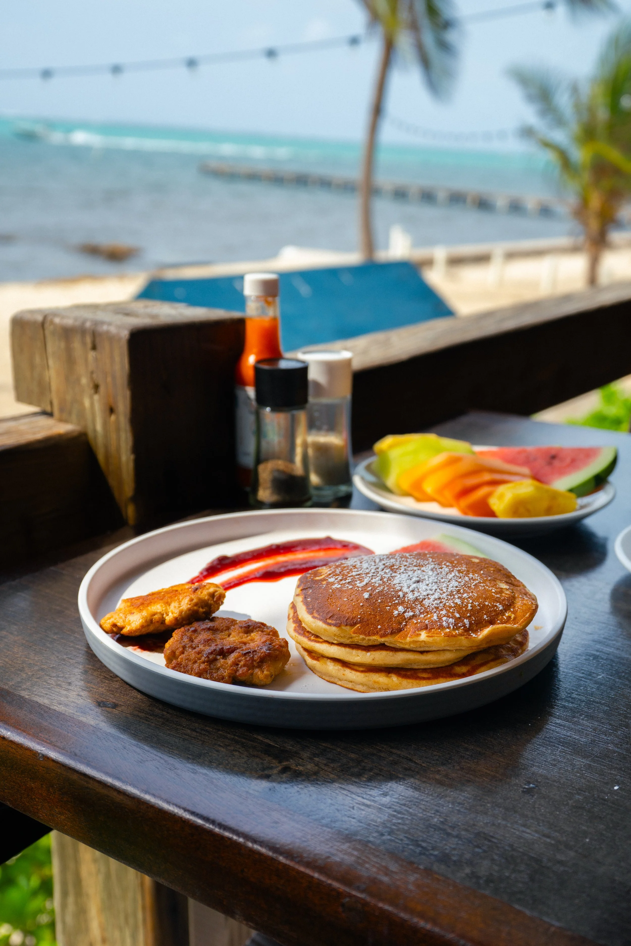 A plate of pancakes with powdered sugar, sausage patties, and syrup, served with a side of fruit slices, at an outdoor seaside location with palm trees and ocean in the background.
