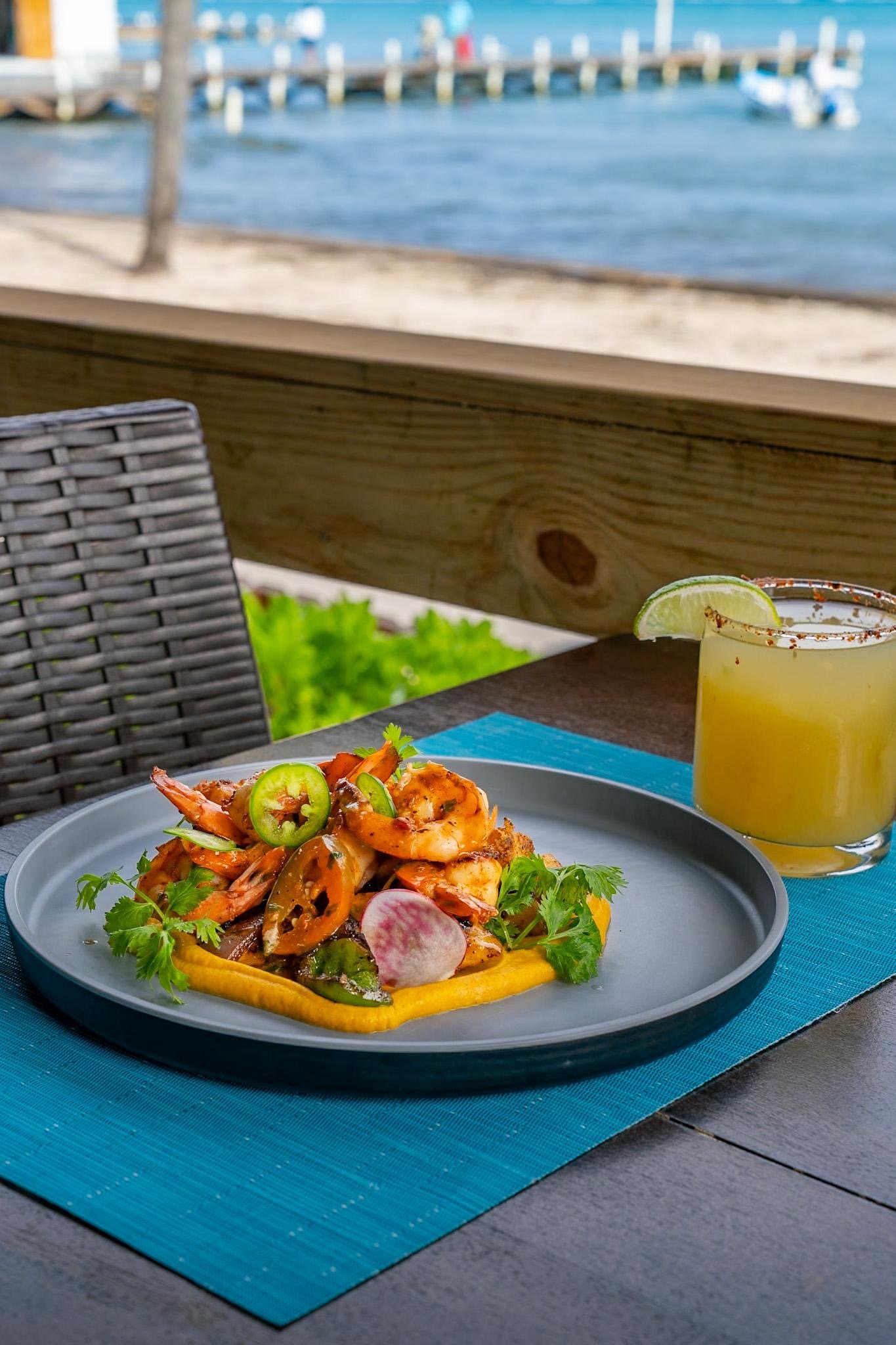A plate of seafood with shrimp, garnished with sliced chili, radish, and cilantro, with a margarita glass topped with a lime wedge in the background, set on a table overlooking the beach.