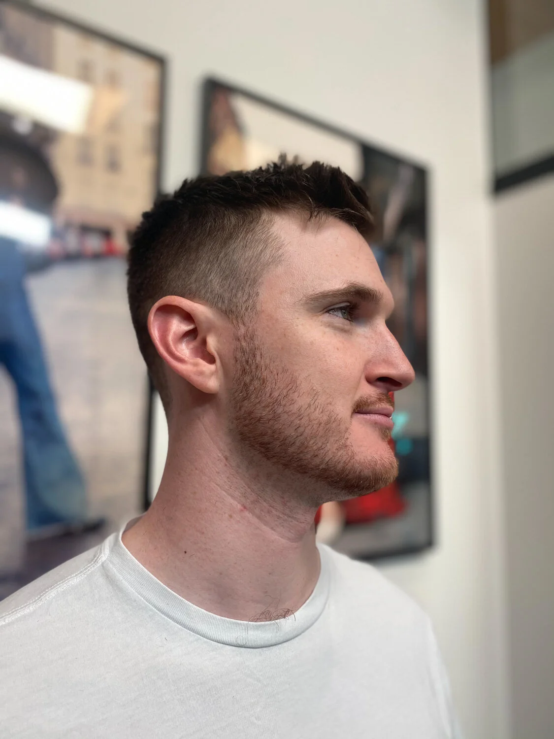 Profile view of a man with short brown hair and a trimmed beard, wearing a white shirt, standing indoors with a blurred background.
