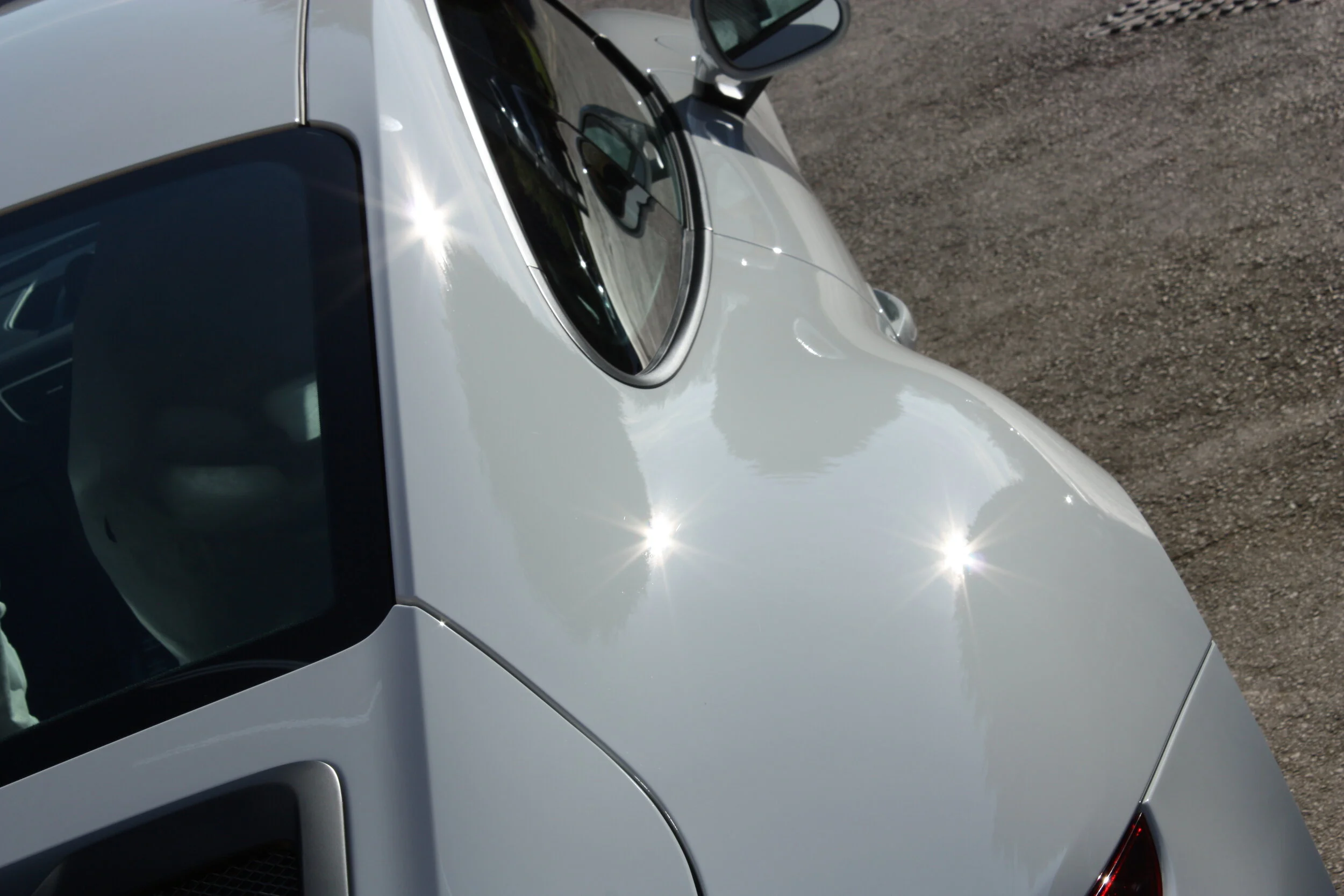 Close-up of a silver sports car's hood, windshield, and driver side window, with reflections of sunlight on the shiny surface, parked on asphalt.