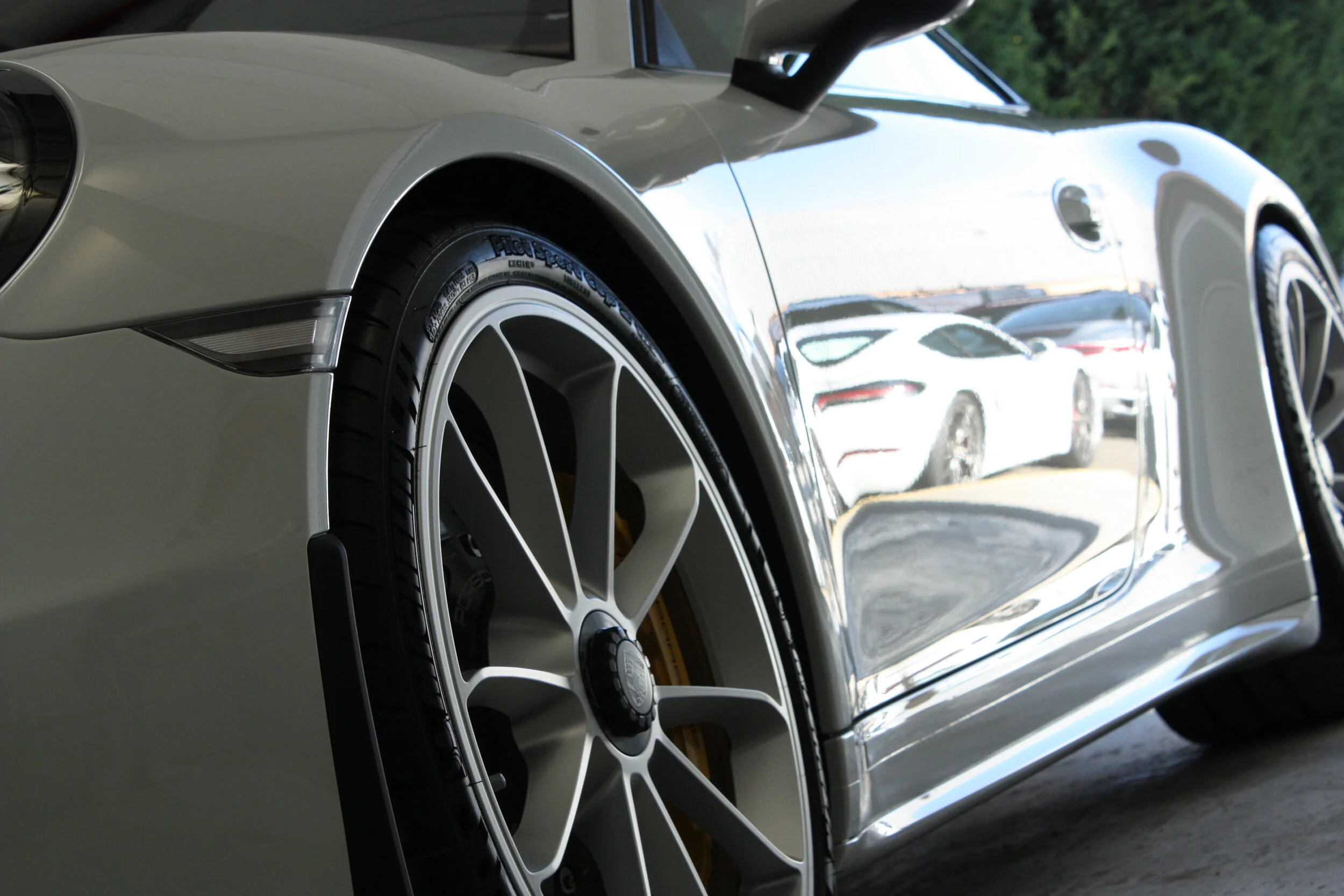 Close-up of a silver luxury sports car front wheel and side mirror, with reflections of other cars in the bodywork.