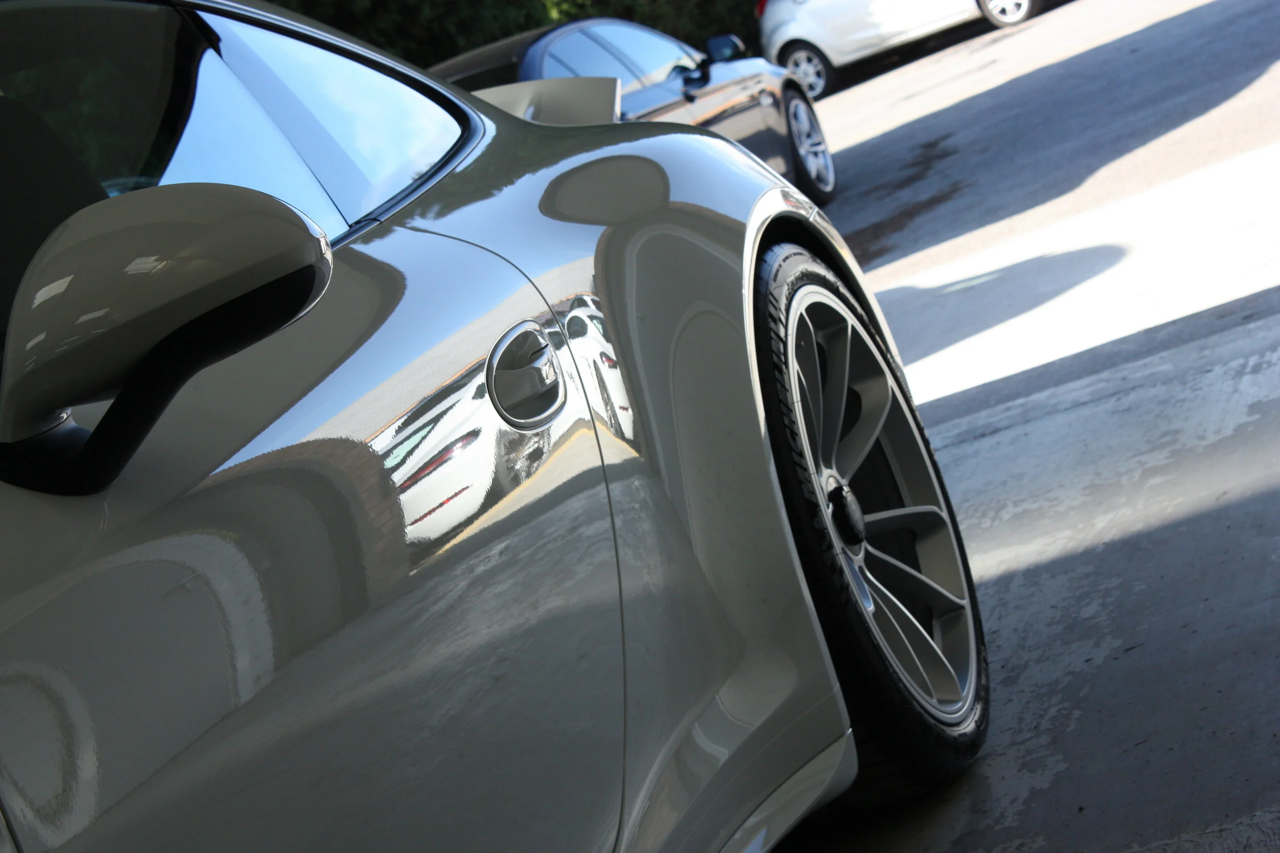 Close-up of a silver sports car with black rims parked in a lot, with other cars in the background.