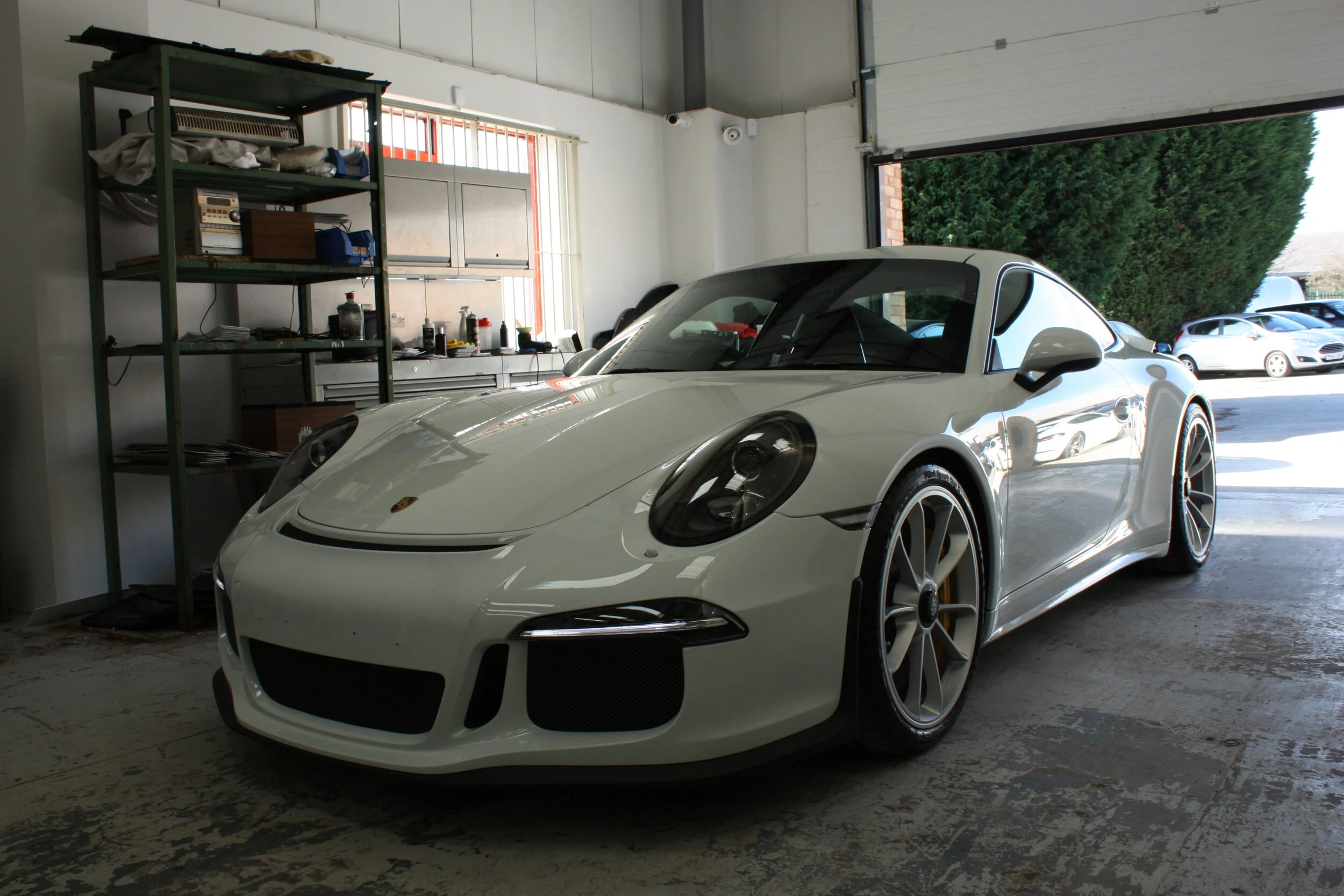 A white Porsche sports car inside a garage with an open door showing parked cars outside.