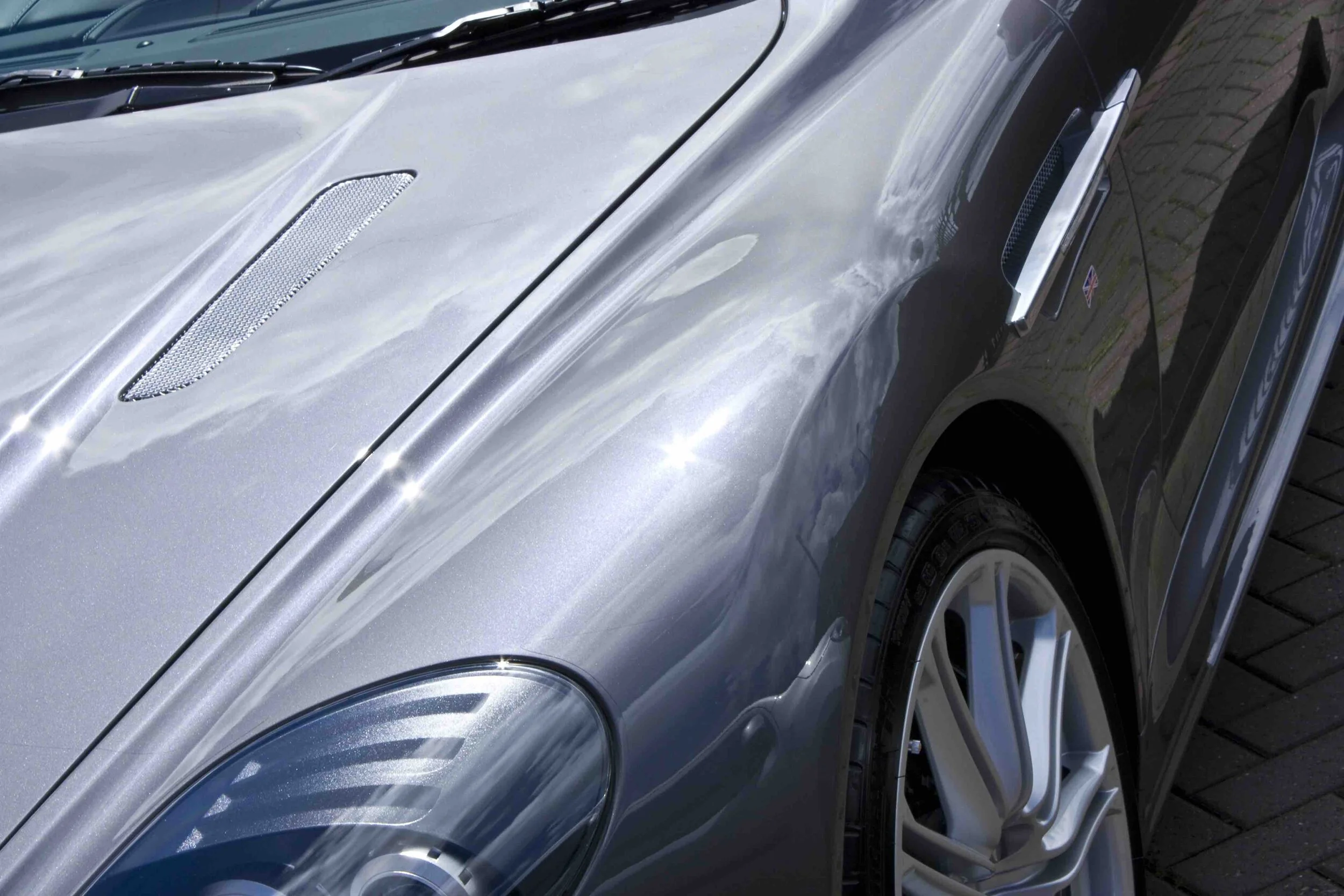 Close-up of a shiny silver sports car with reflections of the sky, clouds, and trees on its hood and fender, showing part of the front wheel and headlight.