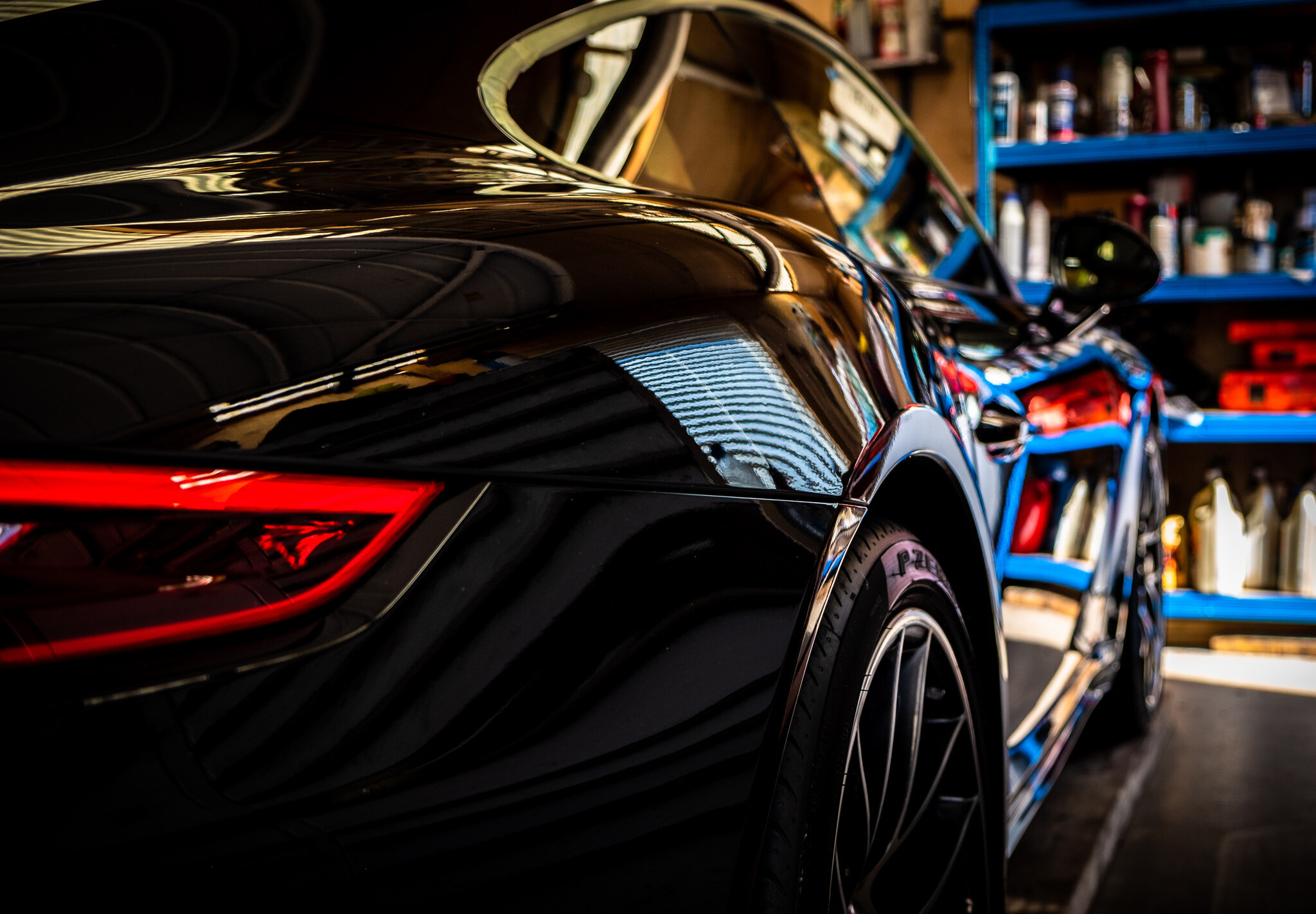 Close-up of a shiny black sports car in a garage, reflecting the surroundings and showing part of the rear and wheel.