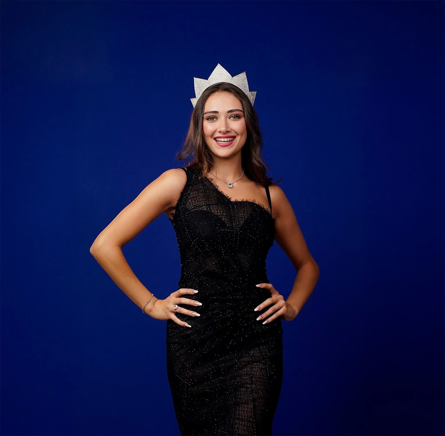 Miss Italia 2025 - Una giovane donna sorridente con una corona di costume, indossa un vestito nero lungo e leggero, posa con le mani sui fianchi su uno sfondo blu.