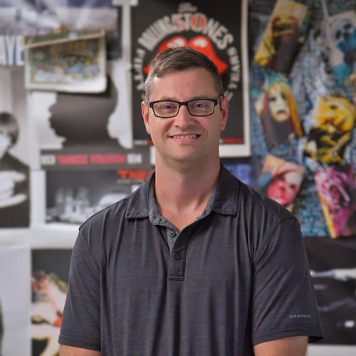A smiling man with glasses standing in front of movie posters and artwork in a gallery or store.