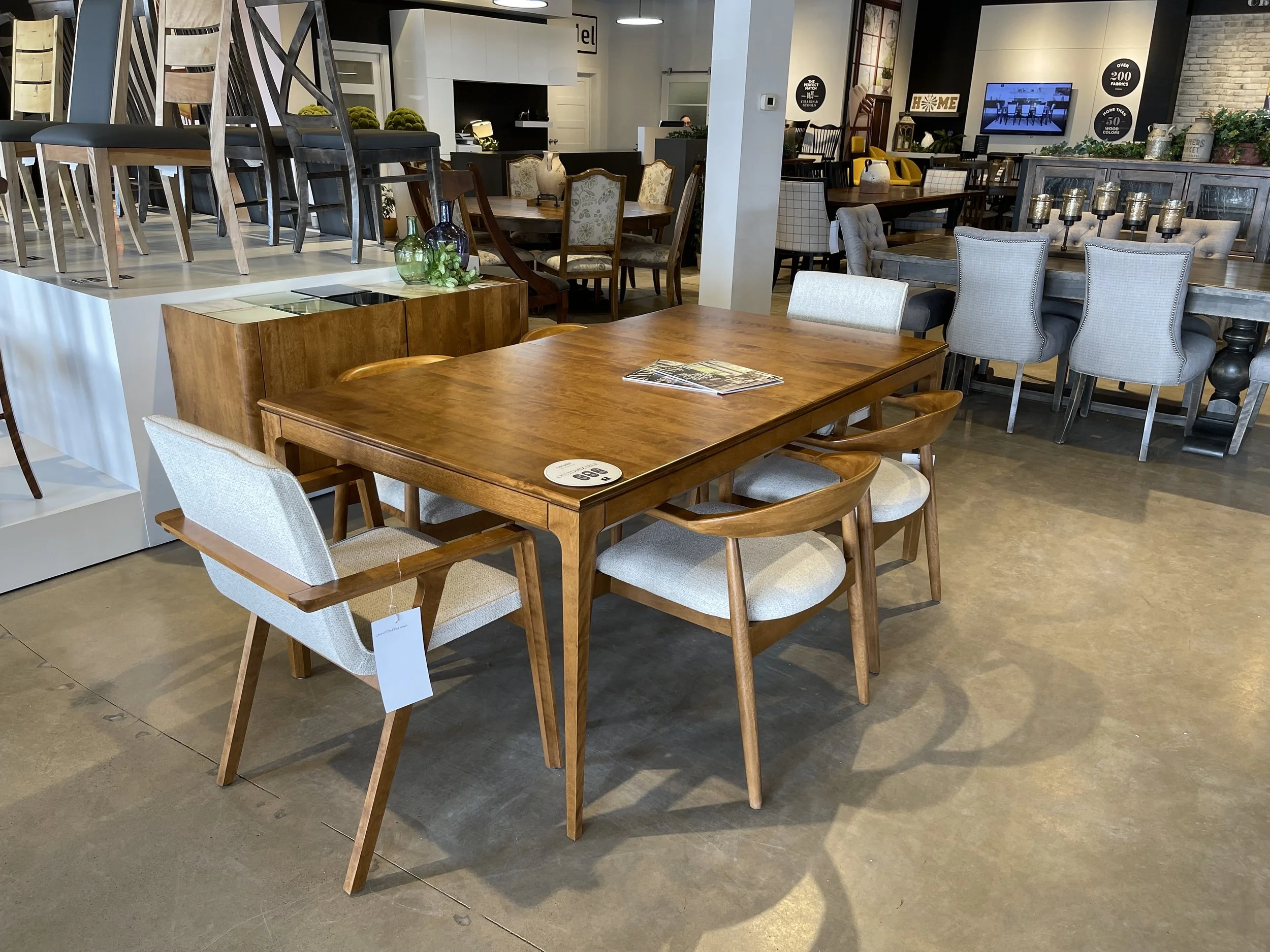 Furniture store display with a wooden dining table, six chairs, and various other tables and chairs in the background.