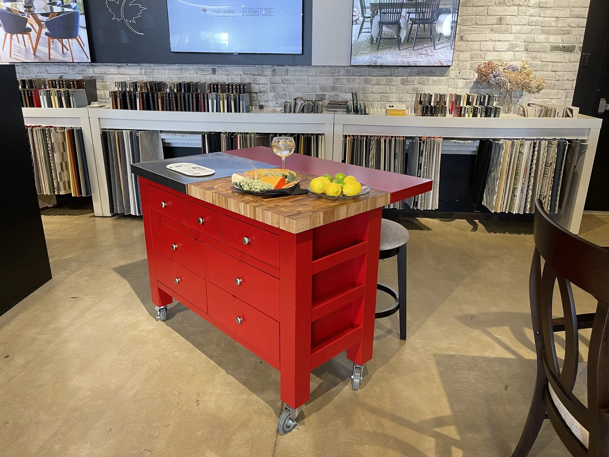 Red kitchen island with a butcher block top, set with a plate of food, a glass of white wine, and a bowl of yellow apples, in a store or showroom with fabric samples and TV screens in the background.