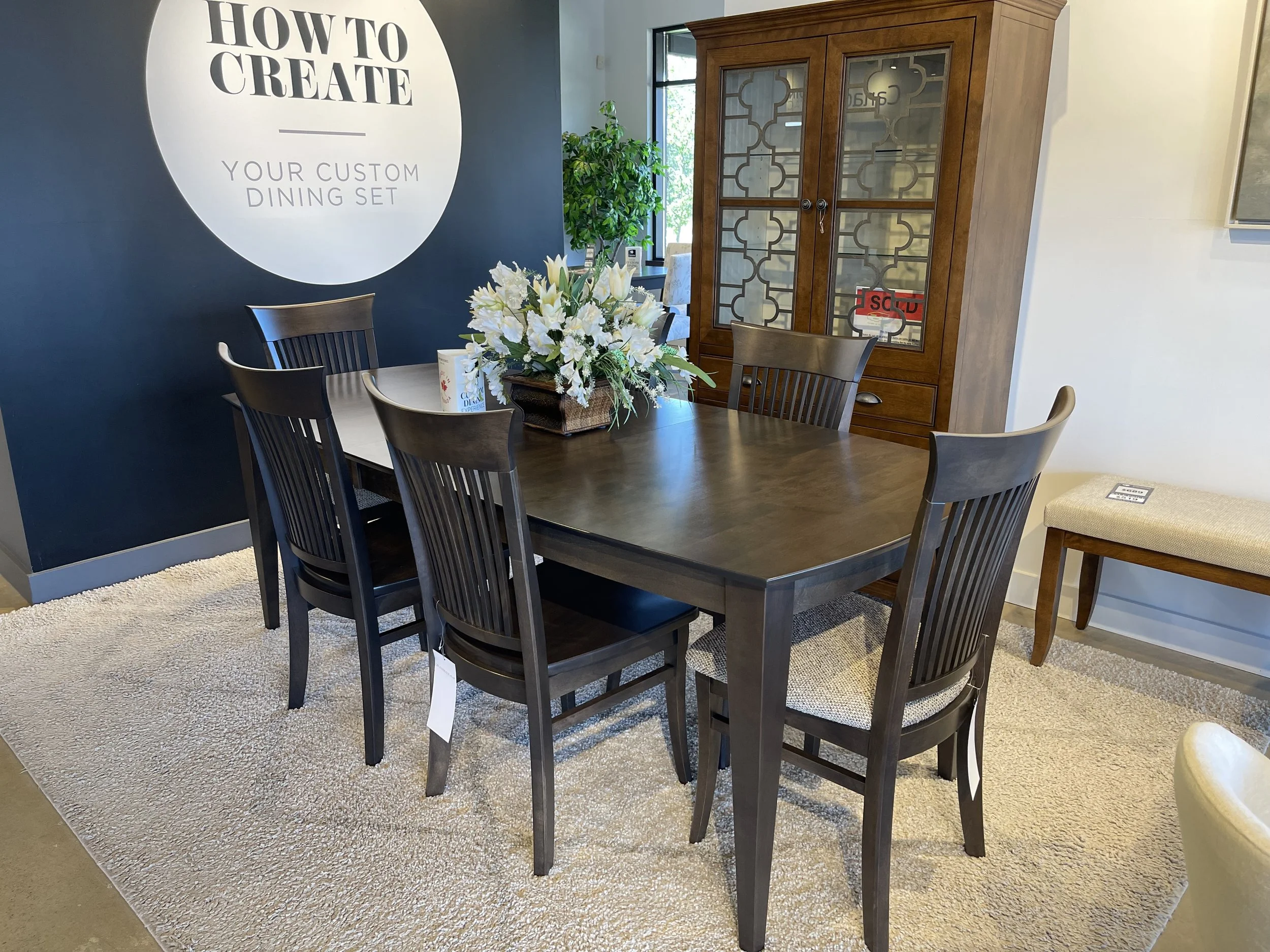 A dining room scene with a dark wooden table and six matching chairs arranged around it. On the table is a large floral centerpiece with white lilies and greenery. A beige cushioned bench with a small tag sits next to the table. Behind the table, the