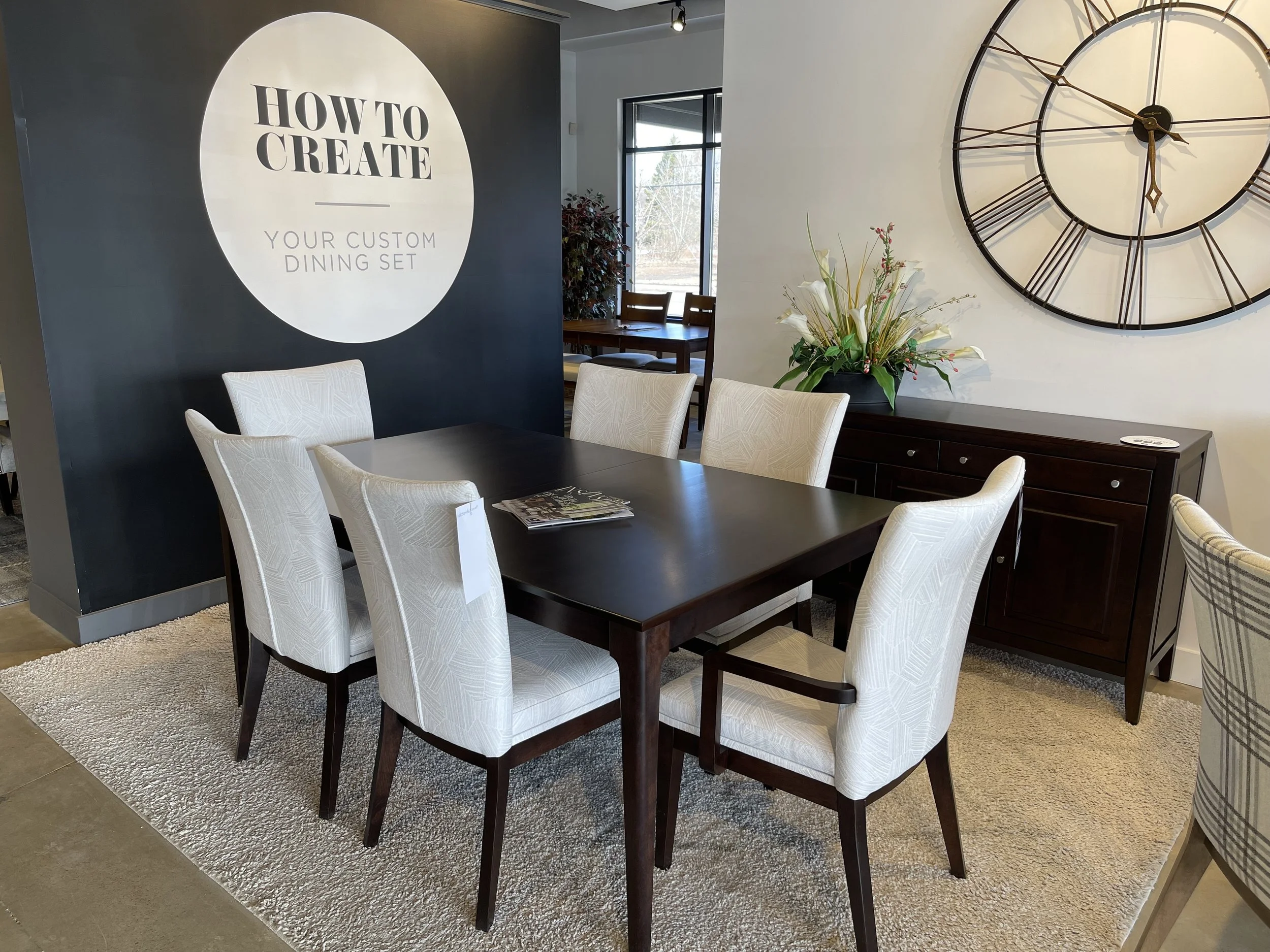 A dining room setup with a black rectangular table surrounded by six white upholstered chairs, a dark wooden sideboard with a flower arrangement, a large wall clock, and a sign on a navy blue wall that reads 'How to Create Your Custom Dining Set.'