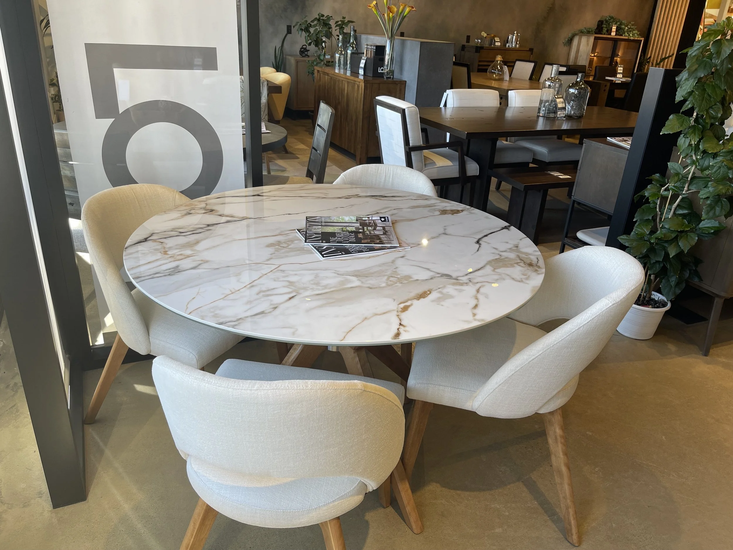 A round marble table with magazines on top, surrounded by four beige upholstered chairs with wooden legs, inside a modern furniture store.