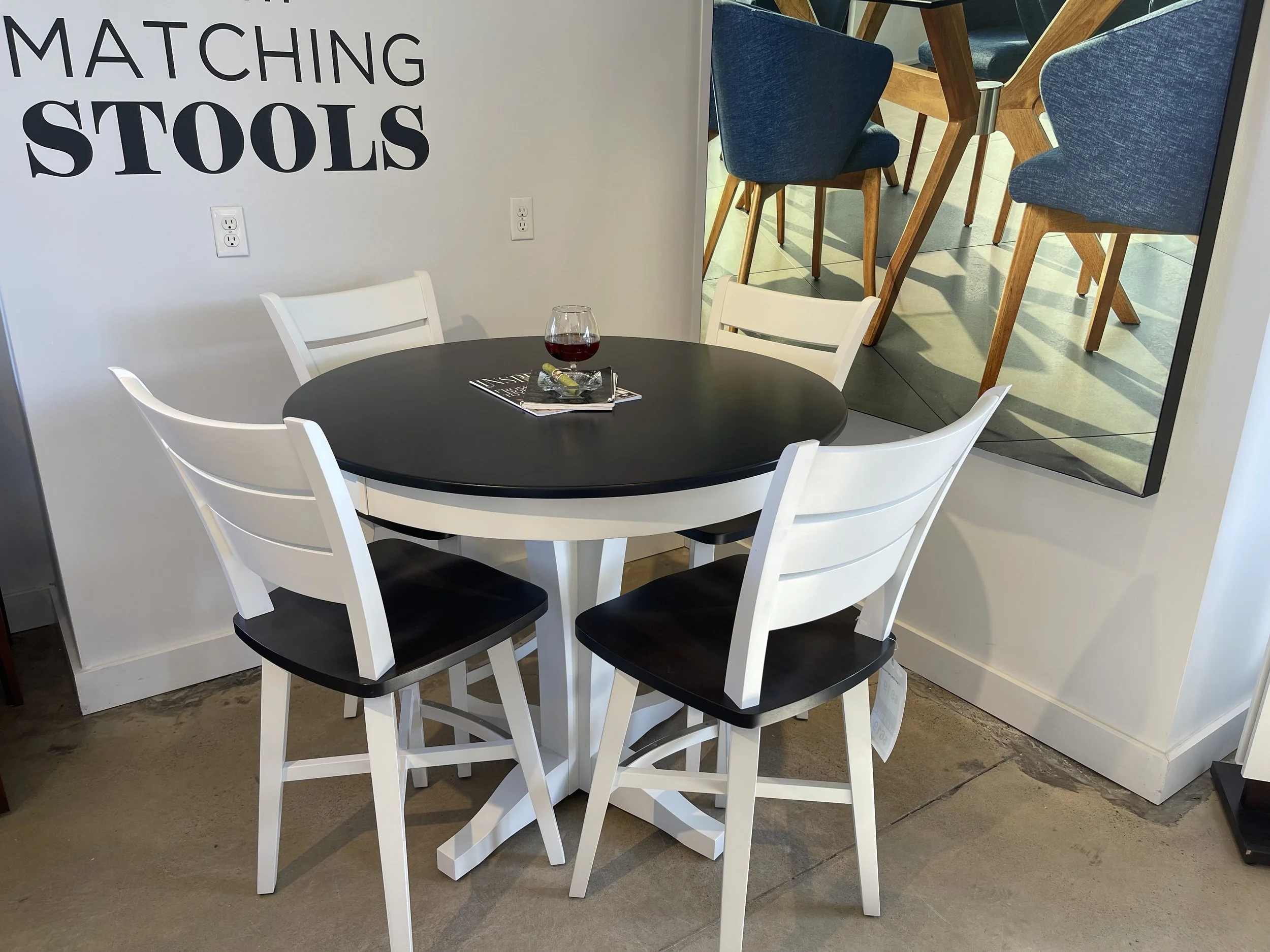 A black round table with four white chairs with black seats. A glass of red wine, a magazine, and a small decorative object are on the table. There is a wall with the words 'Matching Stools' and a mirror reflecting blue chairs and a wooden table.