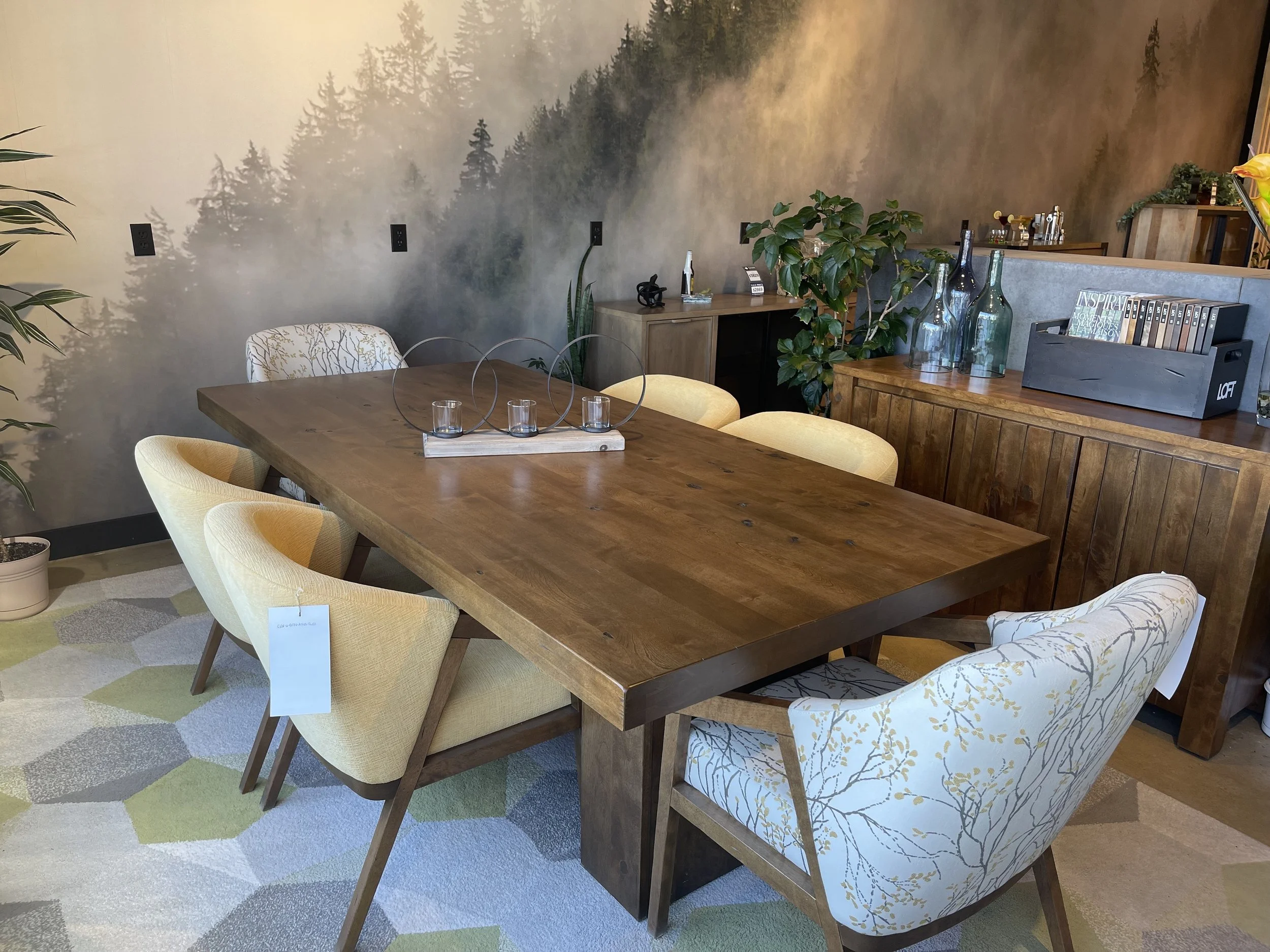A wooden dining table surrounded by six upholstered chairs in a room with a nature-themed mural wall and a wooden sideboard decorated with glass vases and plants.