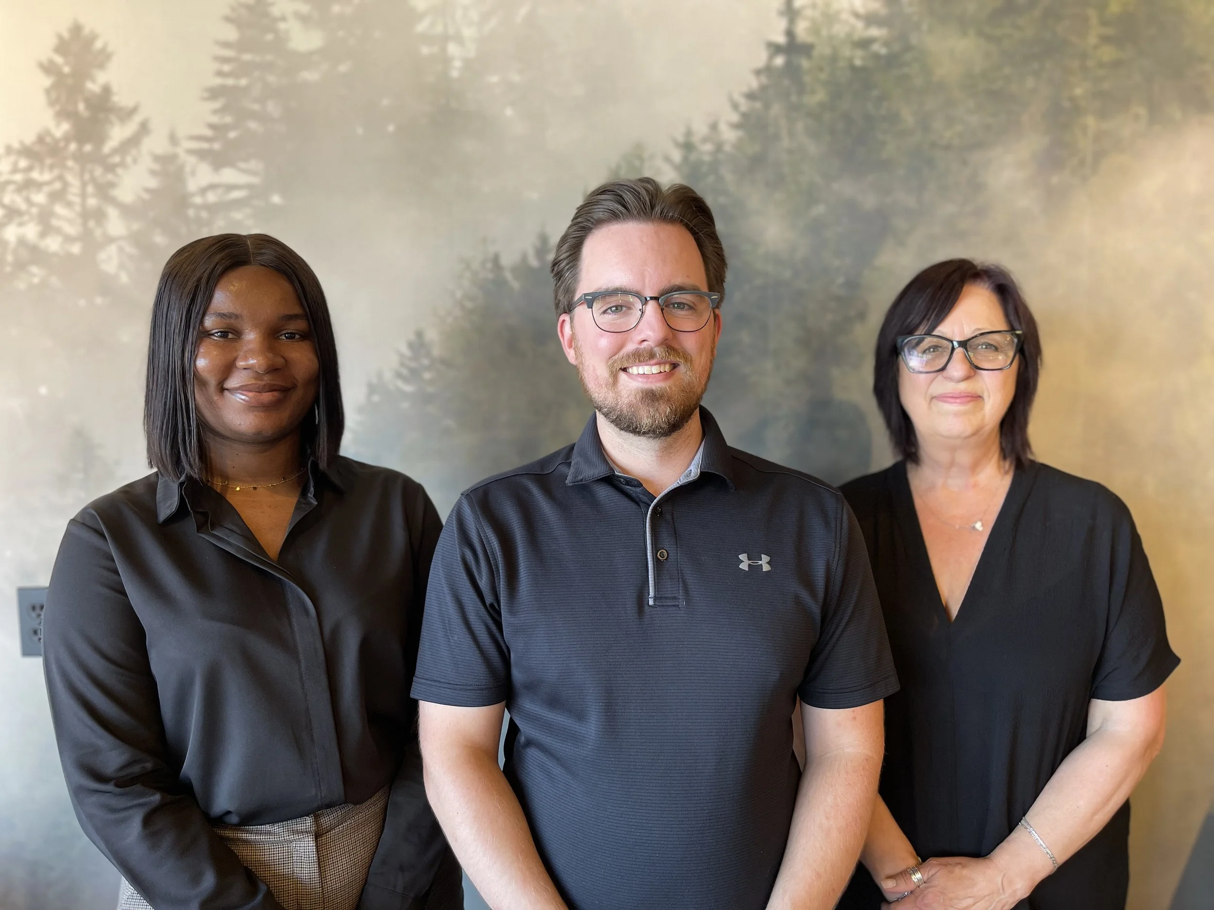 Three professionally dressed individuals, two women and one man, standing in front of a nature-themed mural with trees, smiling at the camera.