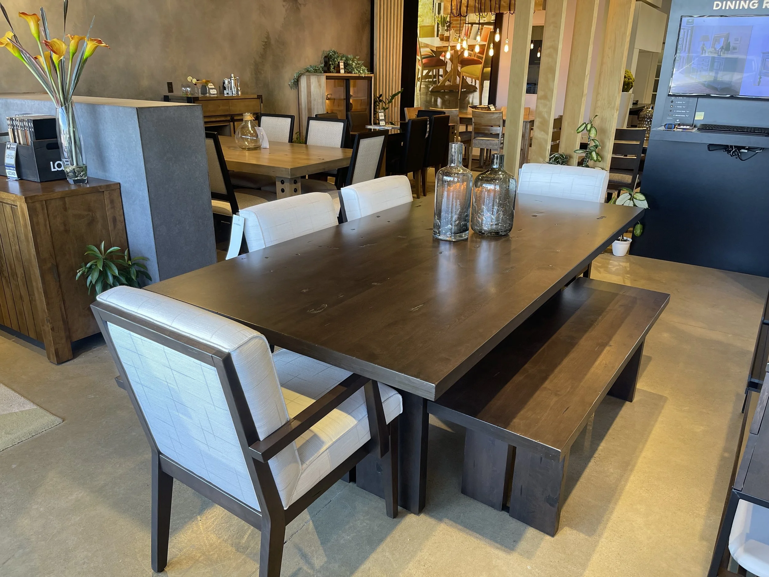 A wooden dining table with white upholstered chairs and a bench, decorated with three large glass vases, inside a modern restaurant or café with additional tables and chairs in the background.