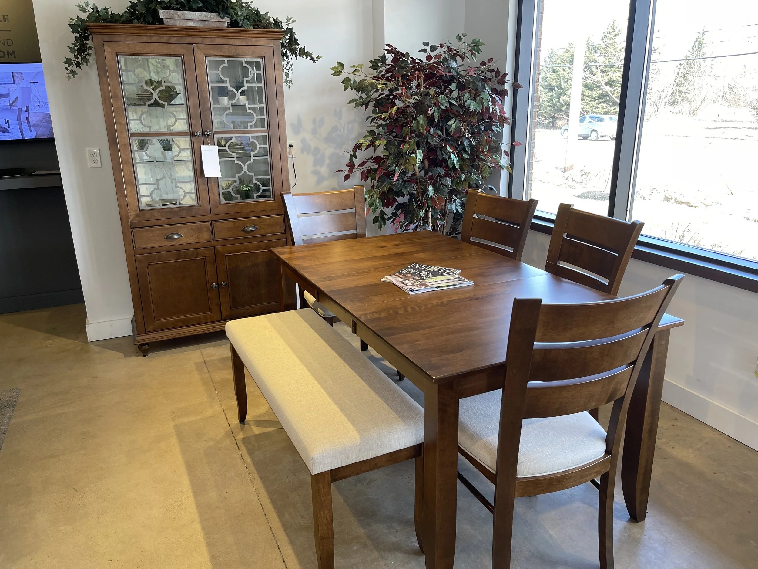 Wooden dining table with four chairs and a bench, near large windows with outdoor view, against a white wall with a large potted plant and a wooden cabinet.