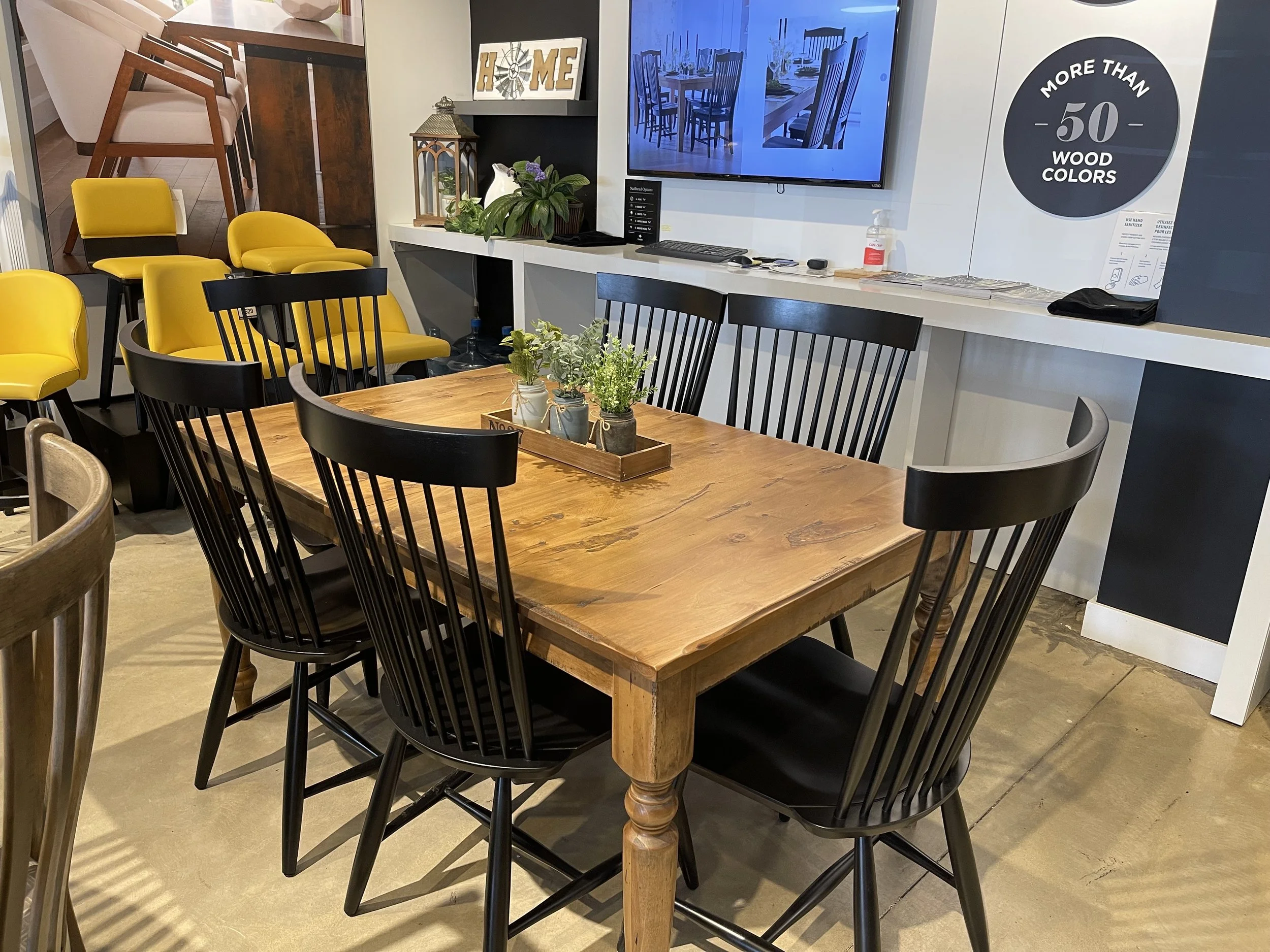 A dining area with a wooden table surrounded by black chairs, small potted plants in the center, and yellow and beige chairs in the background. There is a wall-mounted TV and decorative items on a ledge.