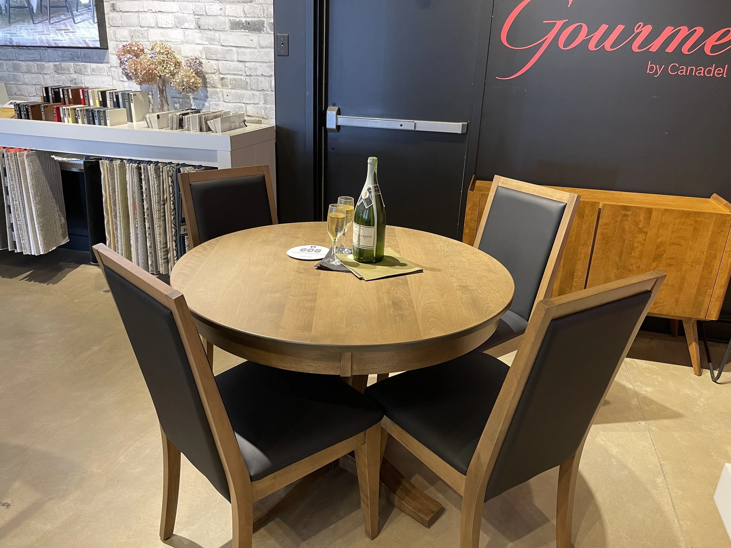 Round wooden table with four black and wood chairs, two glasses of champagne, a bottle of sparkling wine, and a coaster on a green napkin. Background features fabric samples and a black wall with red cursive writing that says "Gourmet by Canadel."