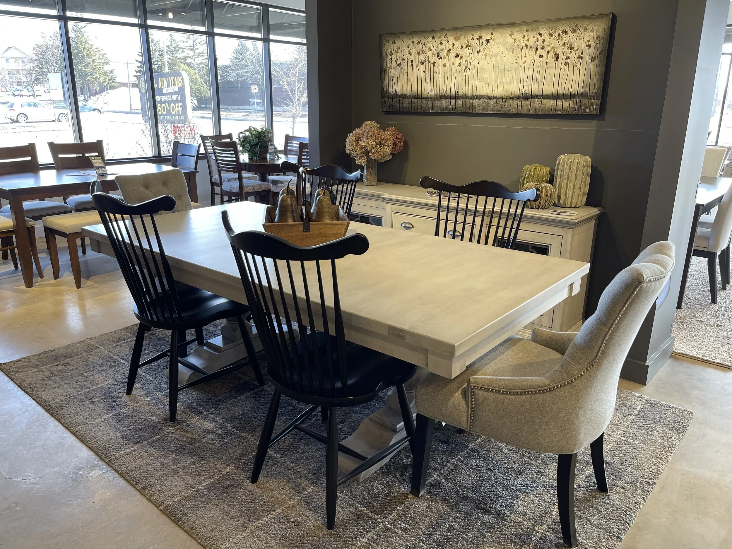 Dining room with a large white table, black and beige chairs, a sideboard with vases, a painting on the wall, and large windows showing a snowy outdoor scene.