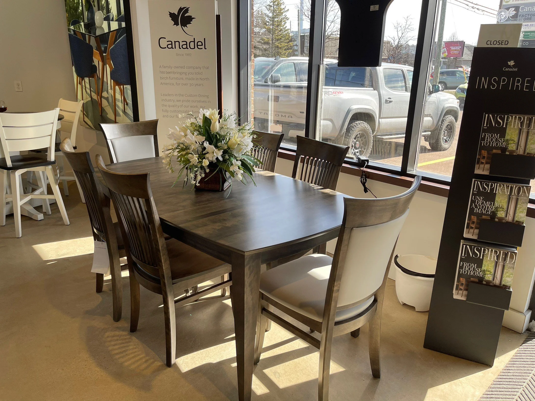 Interior of a furniture store with a dark wood dining table surrounded by various chairs, a vase of white flowers on the table, and promotional materials in the display stand near the window.