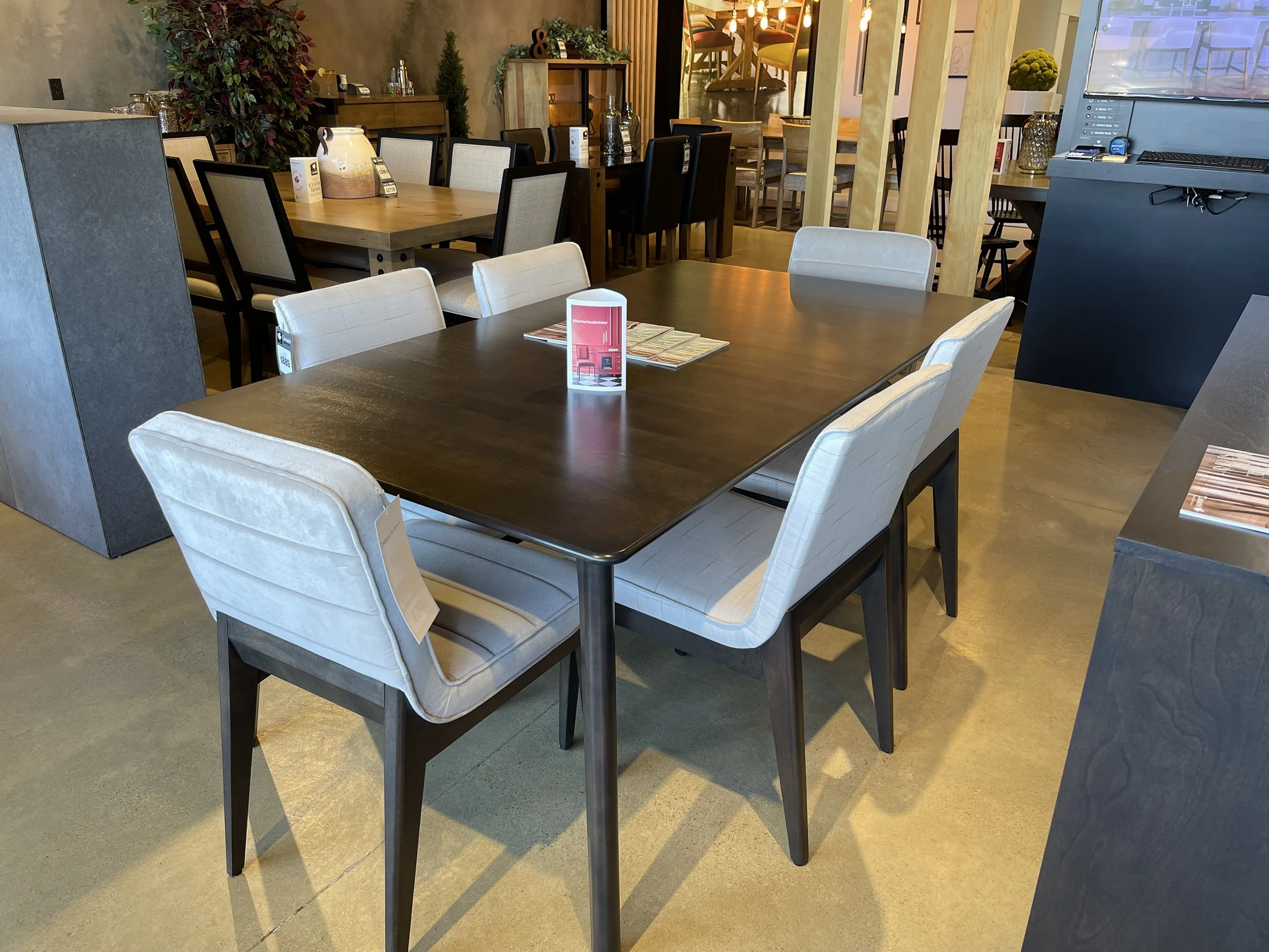 A modern dining area with a dark wood table, white upholstered chairs, and a background of various other tables and chairs in a restaurant setting.