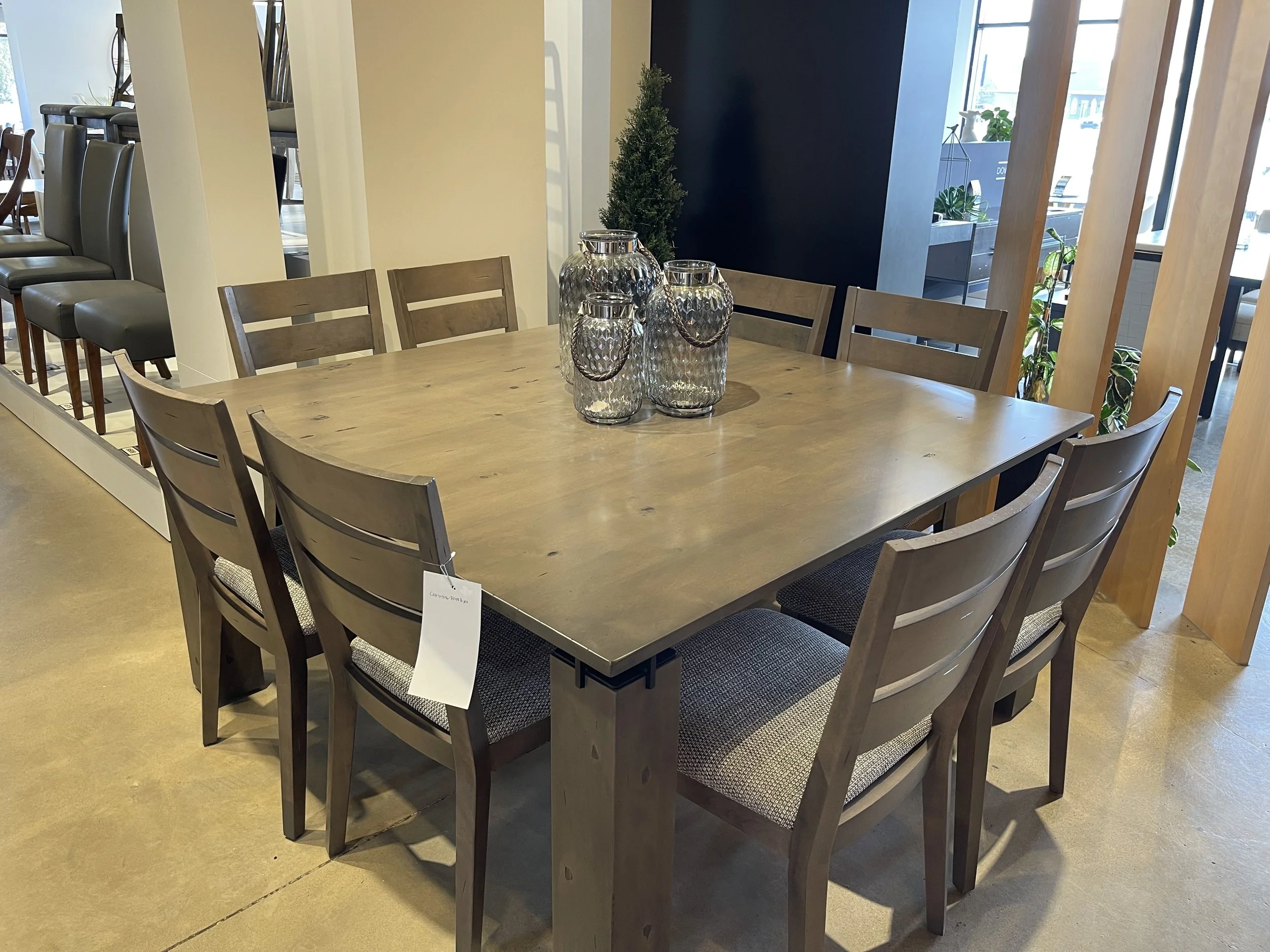 Wooden dining table with six chairs and four decorative glass vases in a modern interior setting with windows and plants.