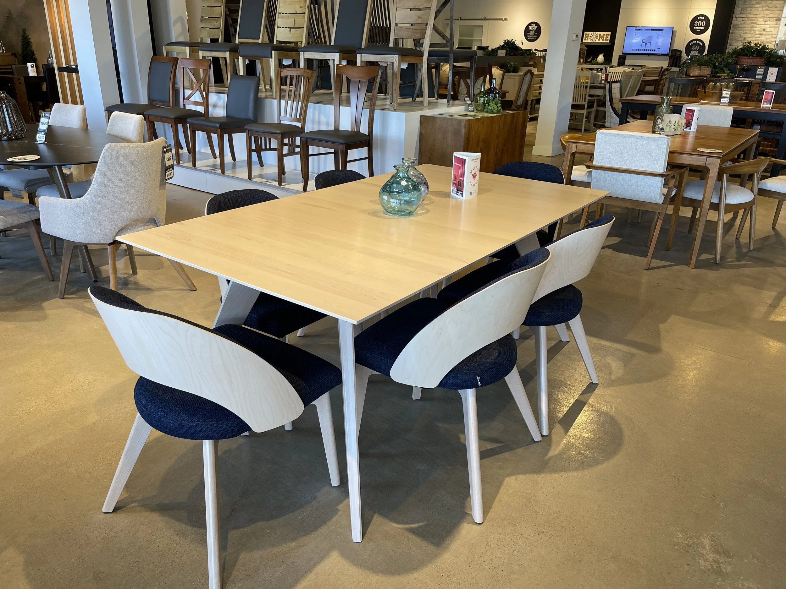 Interior of a furniture store displaying various chairs and tables, some on display shelves and others arranged on the floor, with decorative vases and signs.