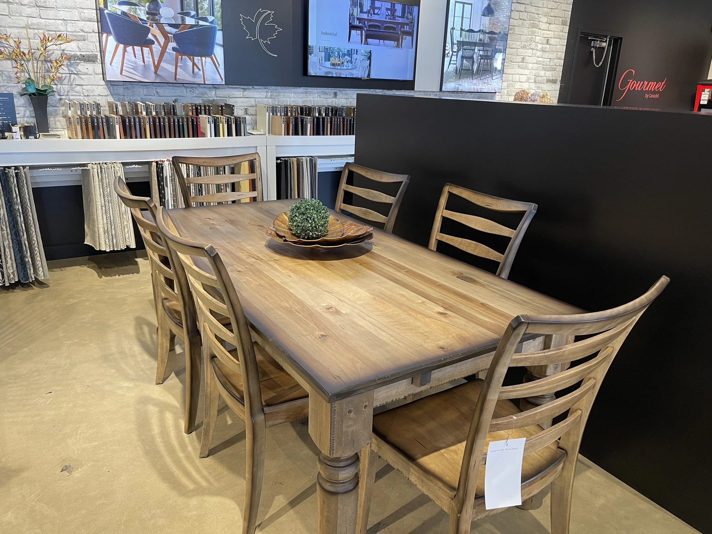 A wooden dining table with six matching chairs, a decorative plate with a small green plant in the center, set in a showroom with fabric samples and large digital screens on the wall.