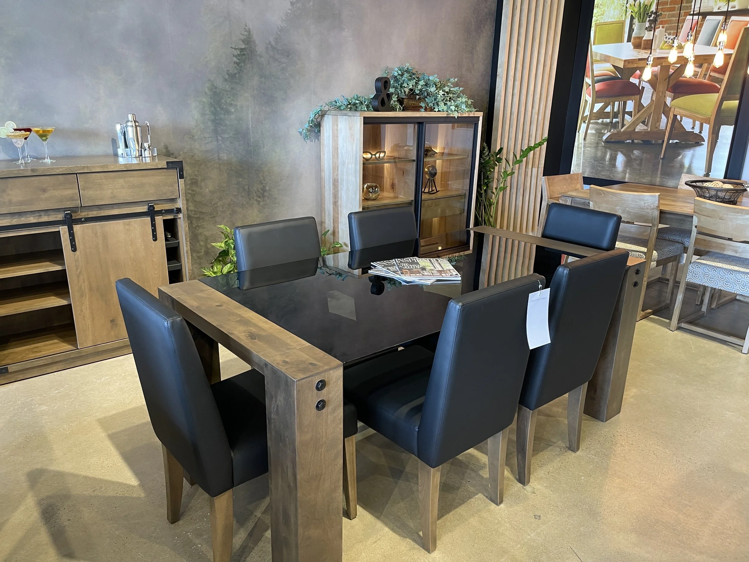 A modern dining room with a black and wood table surrounded by six black leather chairs. There are two wooden sideboards and a glass-front cabinet with decorative items. The room has a mural on one wall and a large window overlooking a patio with out