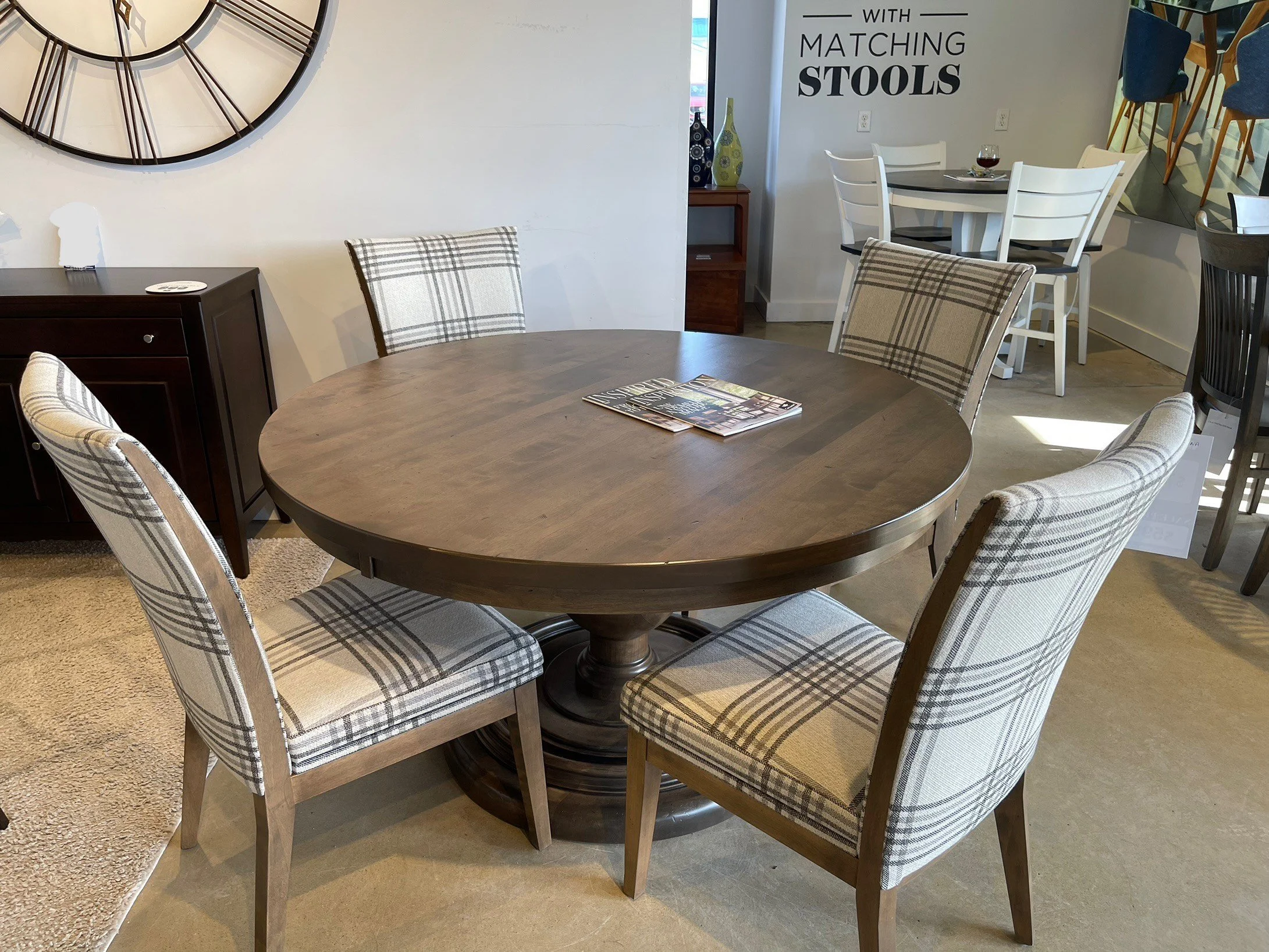 A round wooden dining table with four plaid upholstered chairs arranged around it in a furniture store. The background includes a wall clock, a black cabinet, and a display with more chairs and decorative vases.