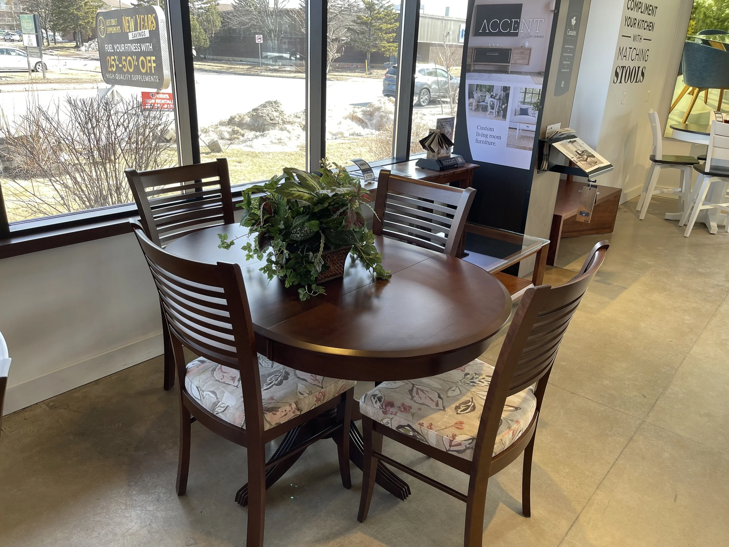 Wooden dining table with four matching chairs and floral cushions, a potted plant centerpiece, inside a store near large windows, with signage and display screens in the background.