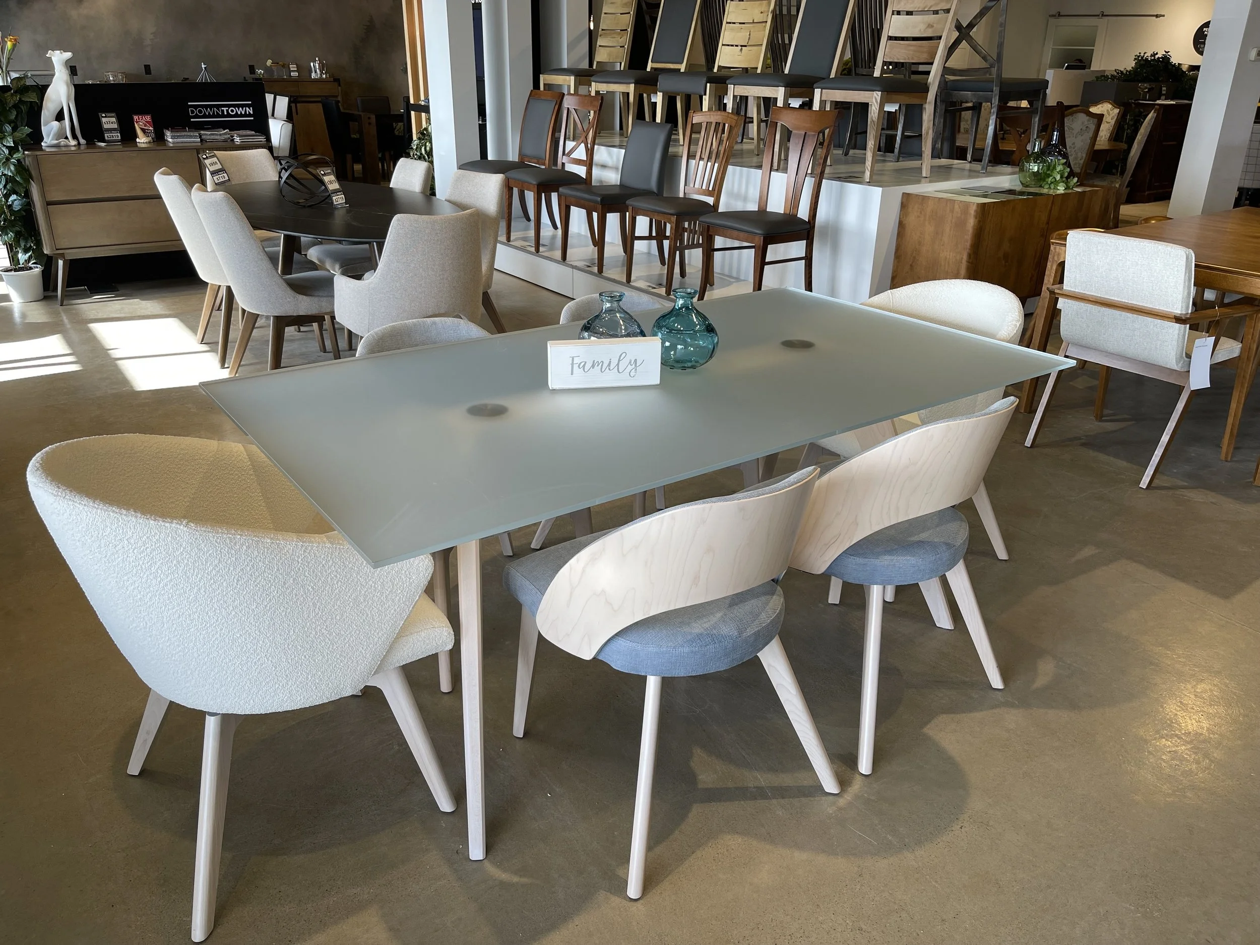 A display of various dining tables and chairs in a furniture store showroom. The focus is on a light-colored, family-sized table with six chairs and a sign that reads 'Family'. The background includes more tables and chairs of different styles and co